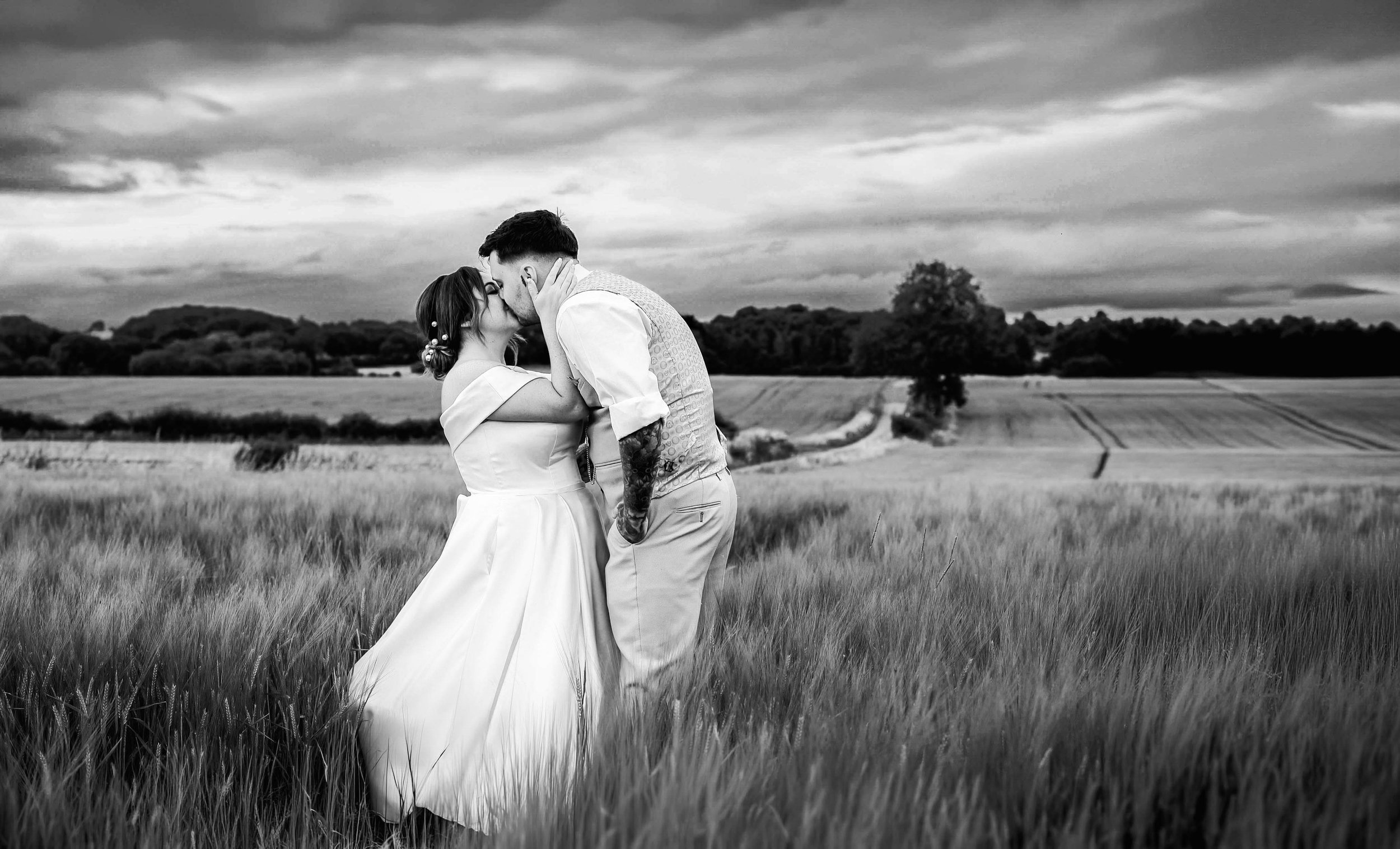 A black and white photo of a couple kissing in a field of tall grass with a scenic landscape and cloudy sky in the background.
