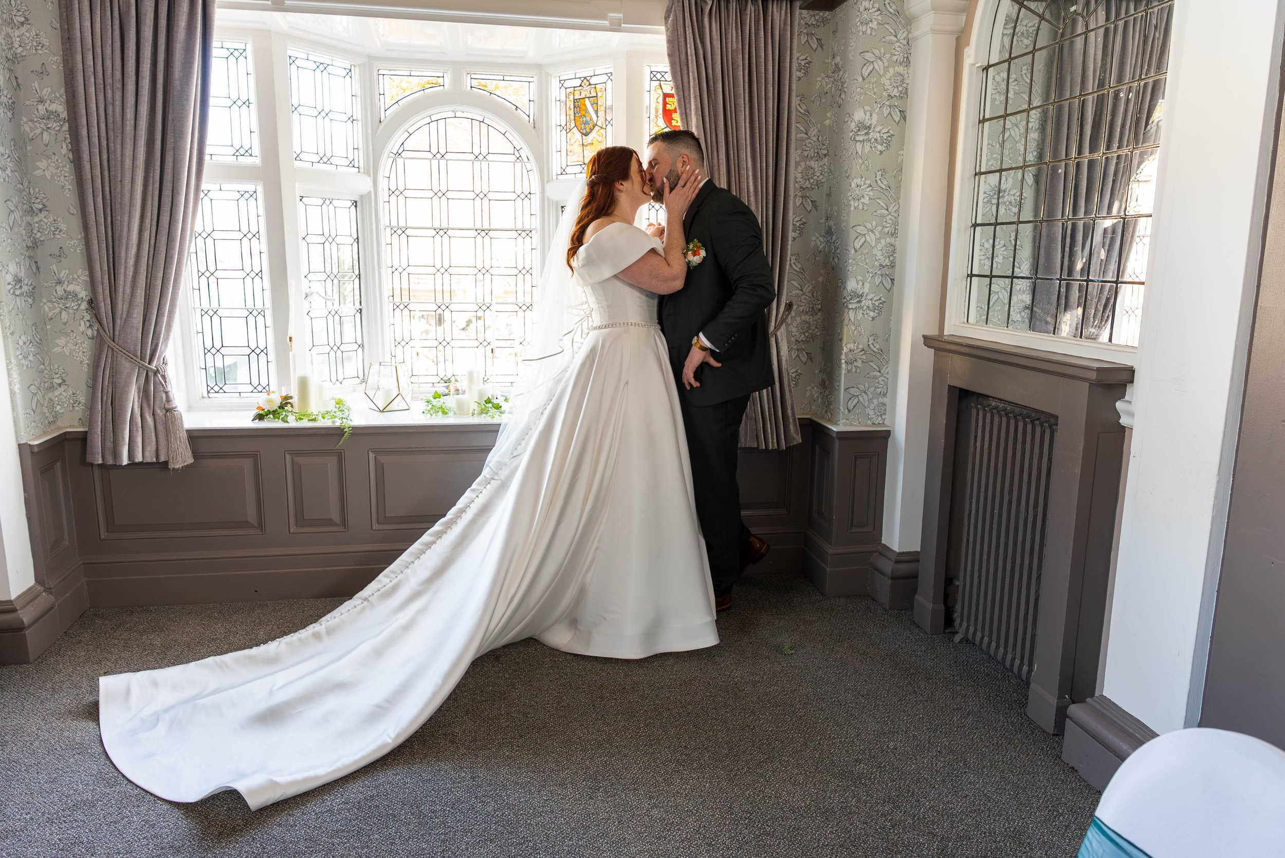 A bride and groom sharing a kiss inside a room with stained glass windows and floral wallpaper, the bride wearing a white wedding gown with a long train and the groom in a black suit.