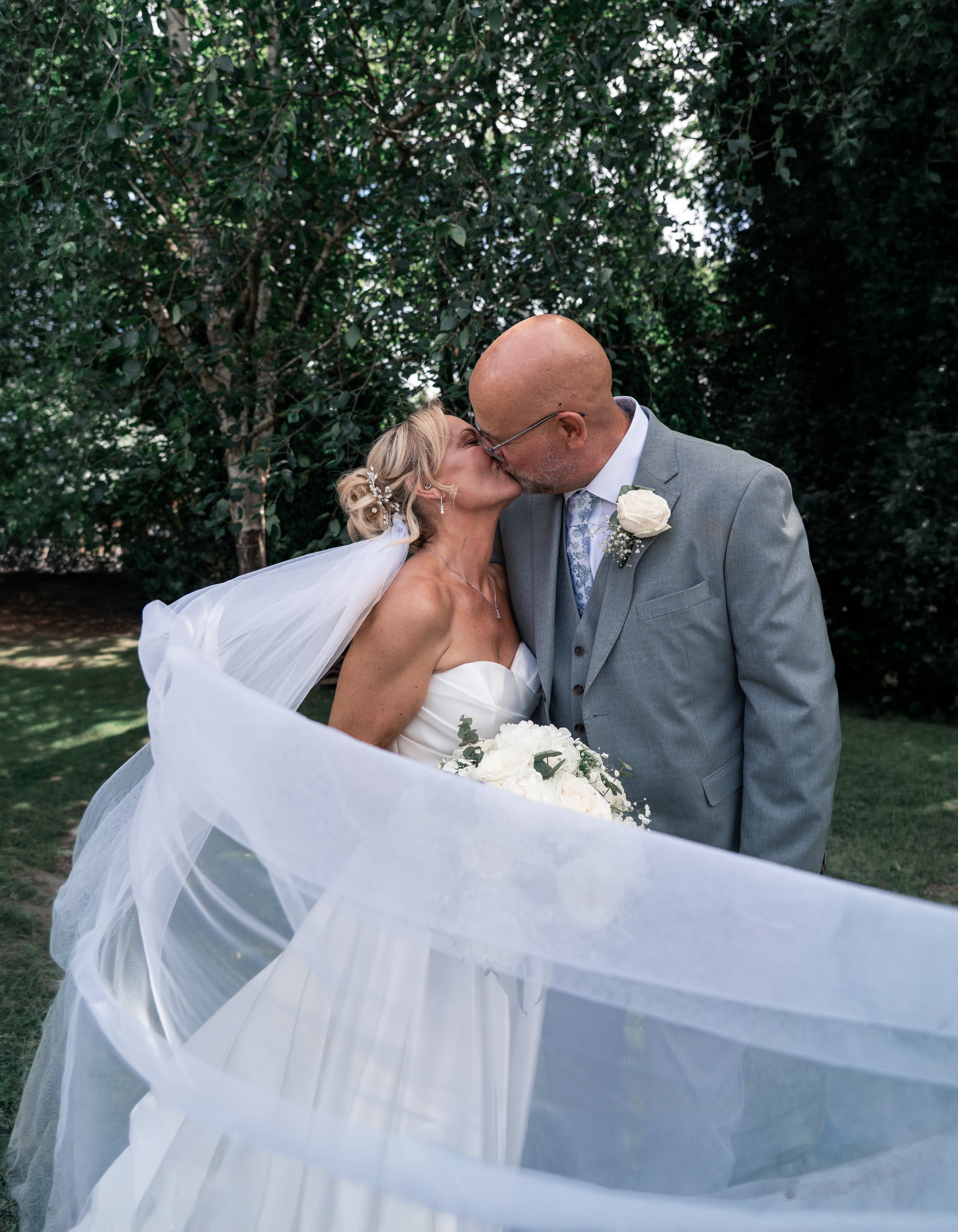 A bride and groom kissing on their wedding day outdoors, surrounded by trees.