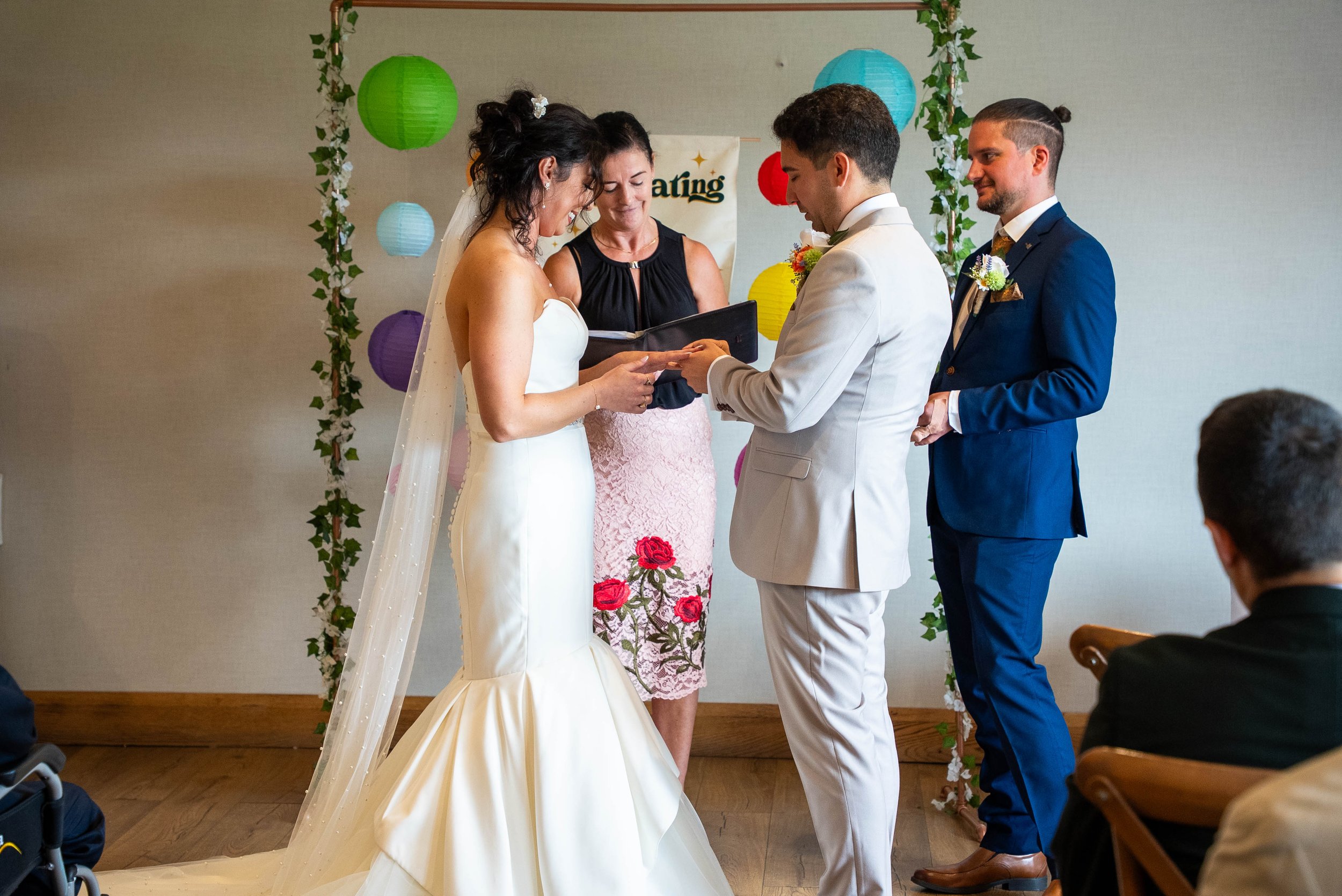 A wedding ceremony with a bride and groom exchanging vows, officiant woman reading from a book, and best man standing behind, decorated with colorful paper lanterns and floral garlands.