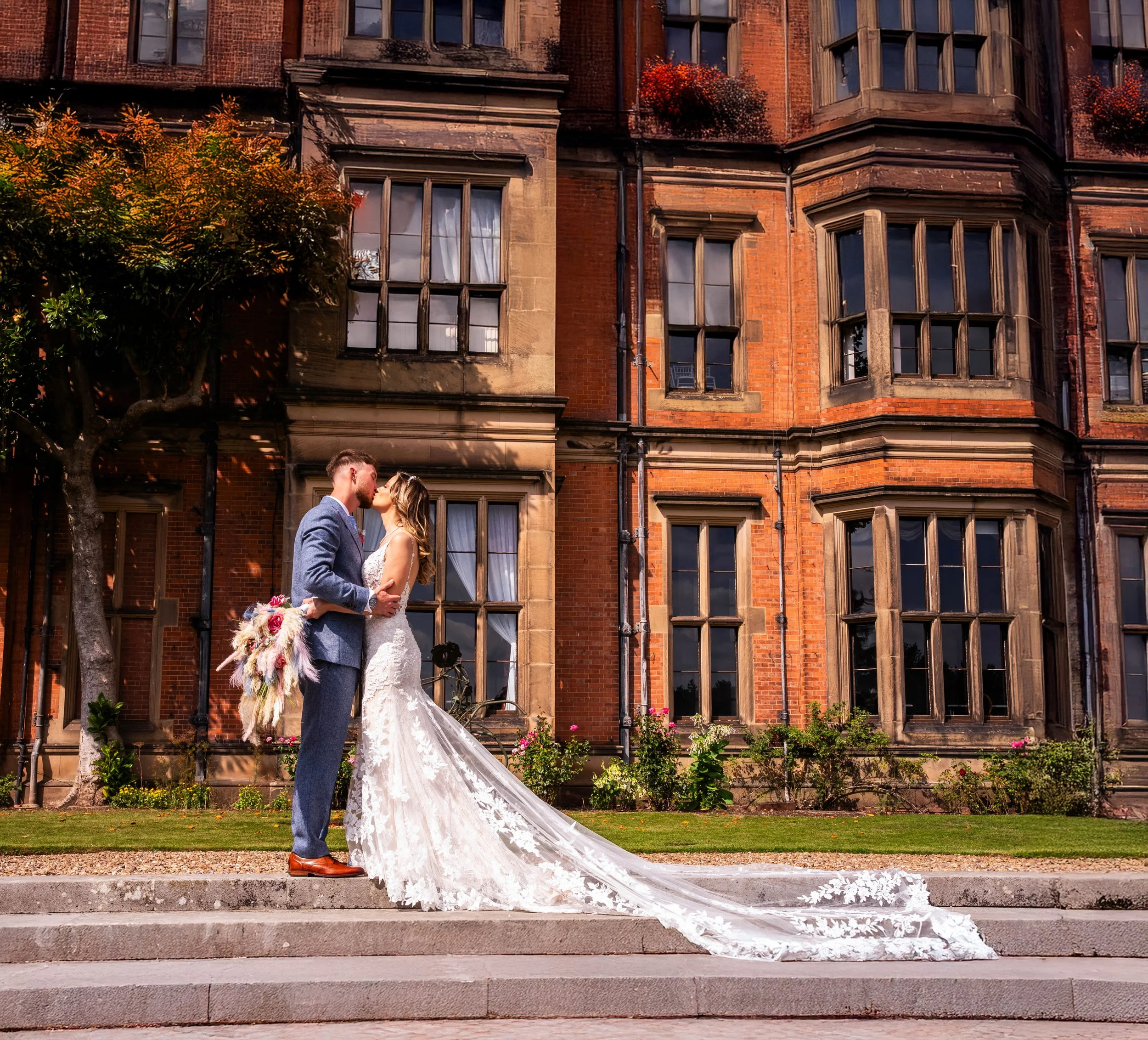 A bride and groom kissing on steps in front of a brick building, with the bride in a long lace wedding gown and the groom in a blue suit holding a bouquet.