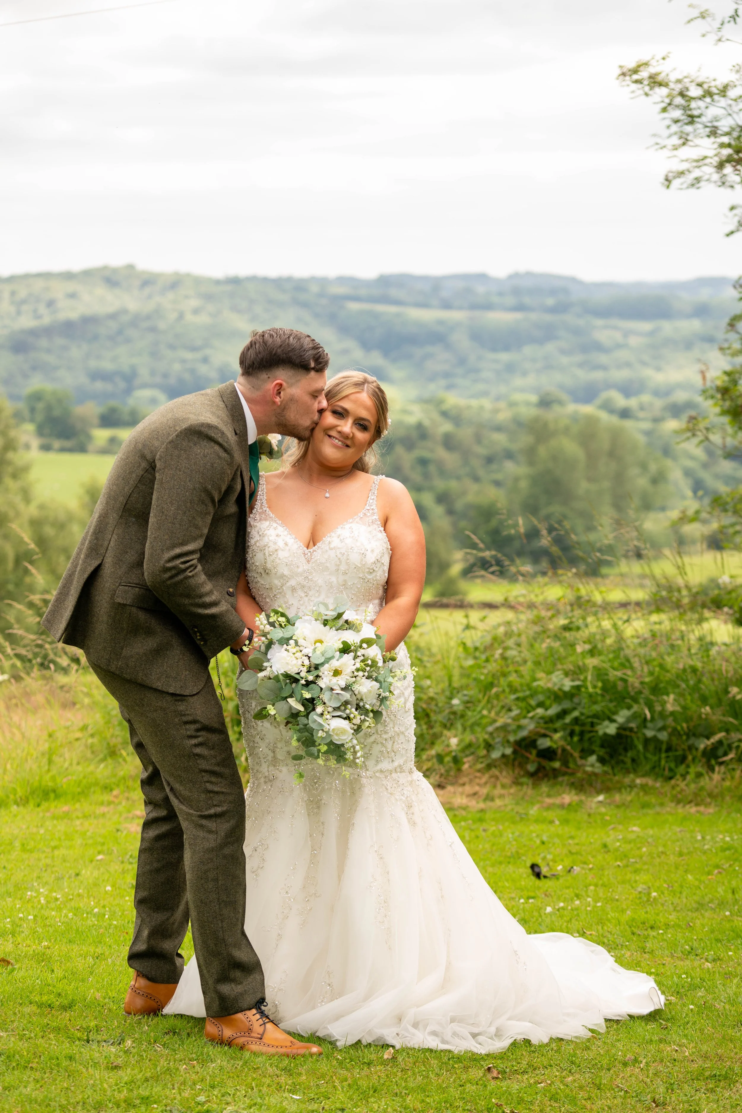 A newlywed couple in wedding attire standing on a grassy field with rolling hills and trees in the background. The groom is kissing the bride on the cheek, and she is holding a bouquet of white flowers and greenery. The bride is wearing a white lace 