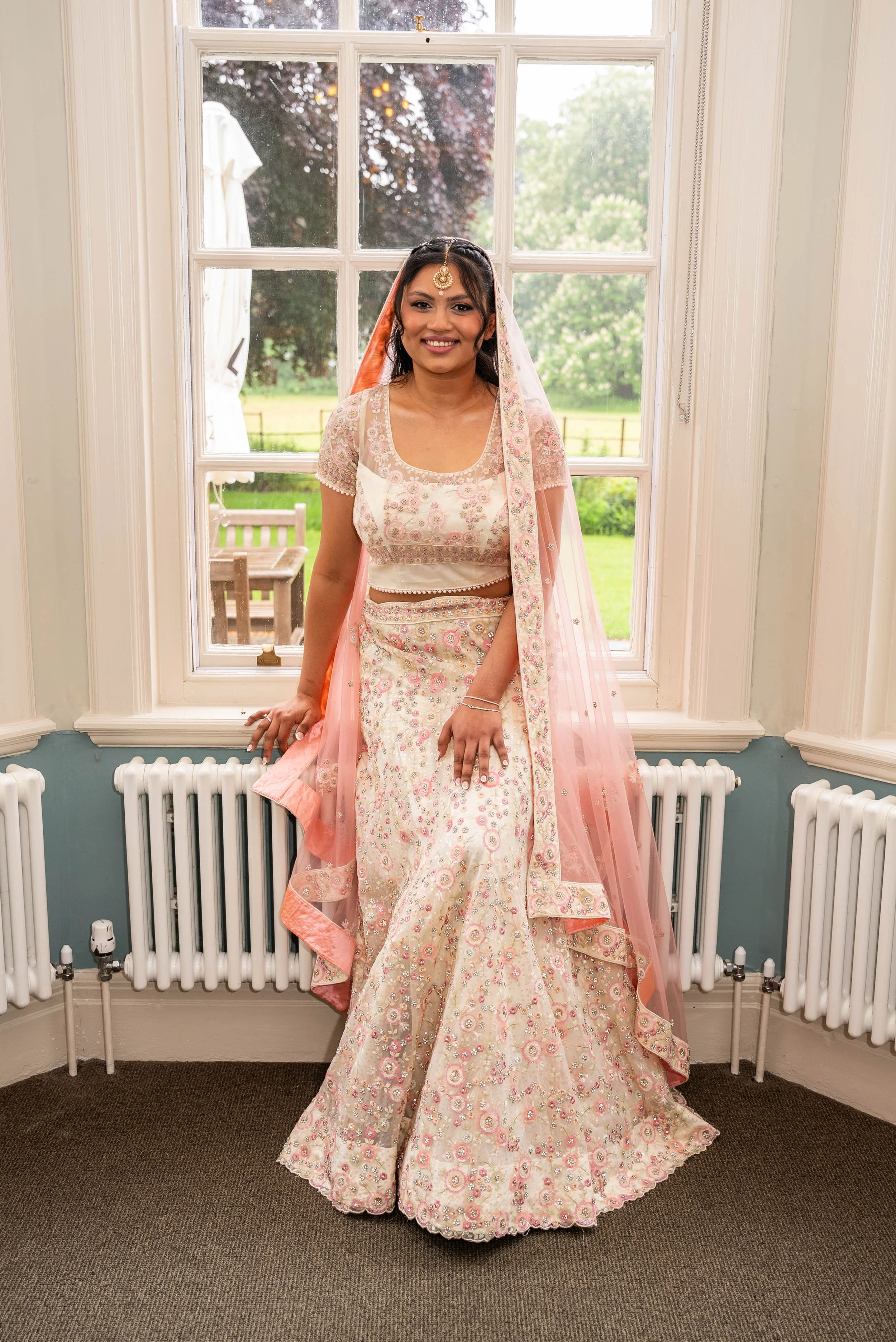 A woman dressed in traditional South Asian bridal attire standing indoors near a large window with green outdoor scenery in the background.