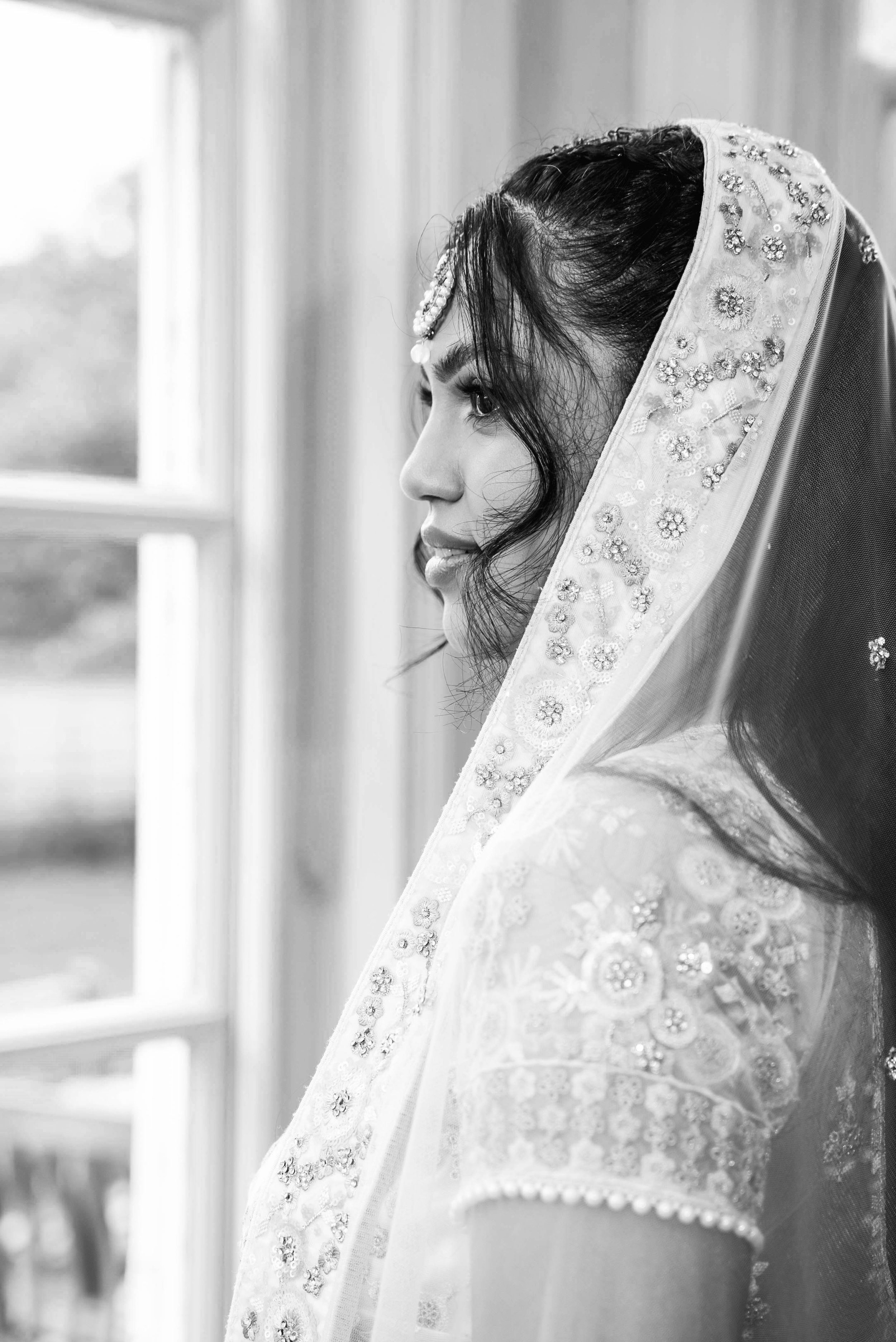 Black and white photo of a woman wearing a lace veil with floral embroidery and beaded embellishments, looking out a window.