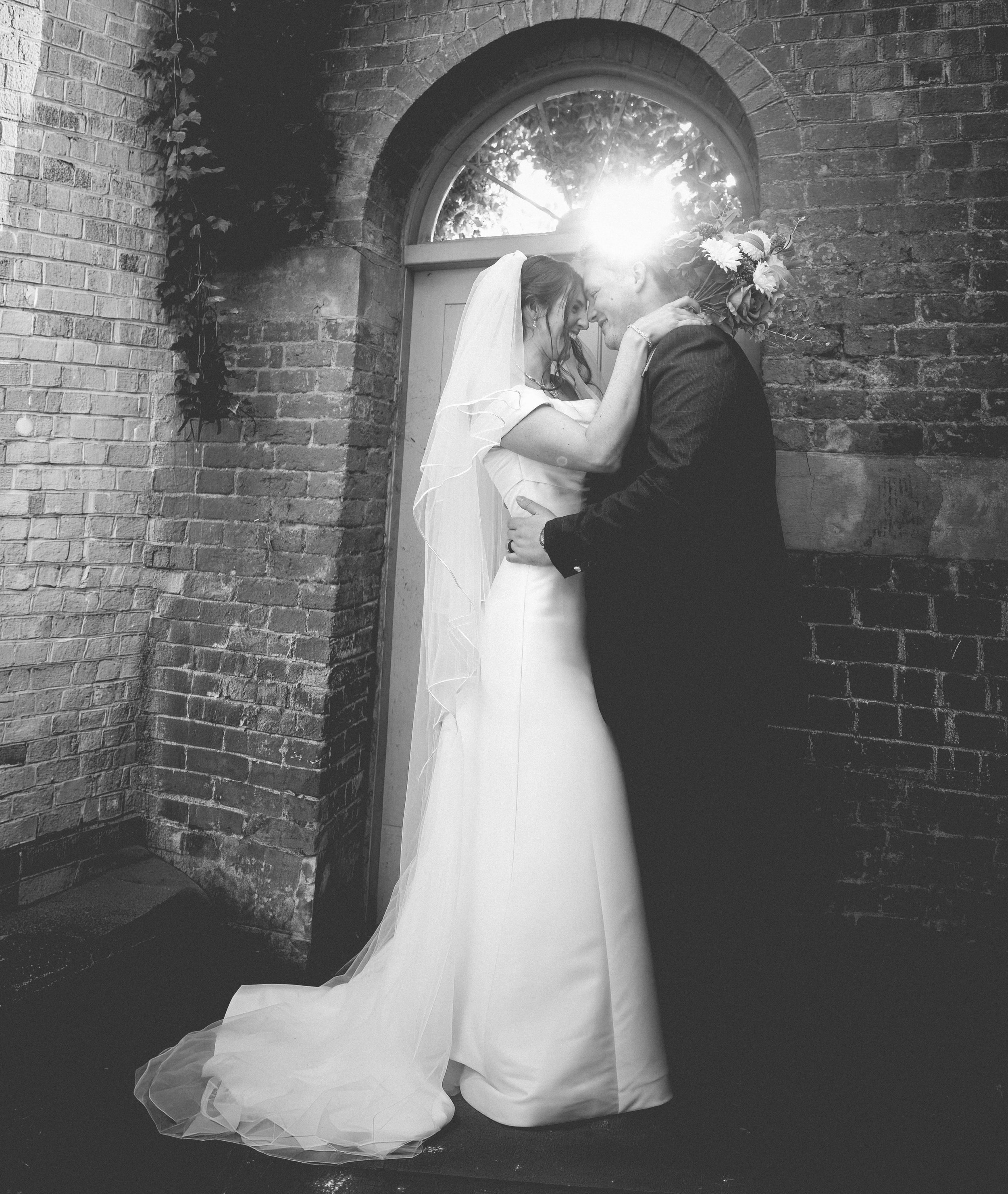 A bride and groom smiling and holding each other closely in front of a brick wall with an arched window, sunlight shining through the window, black and white photo.