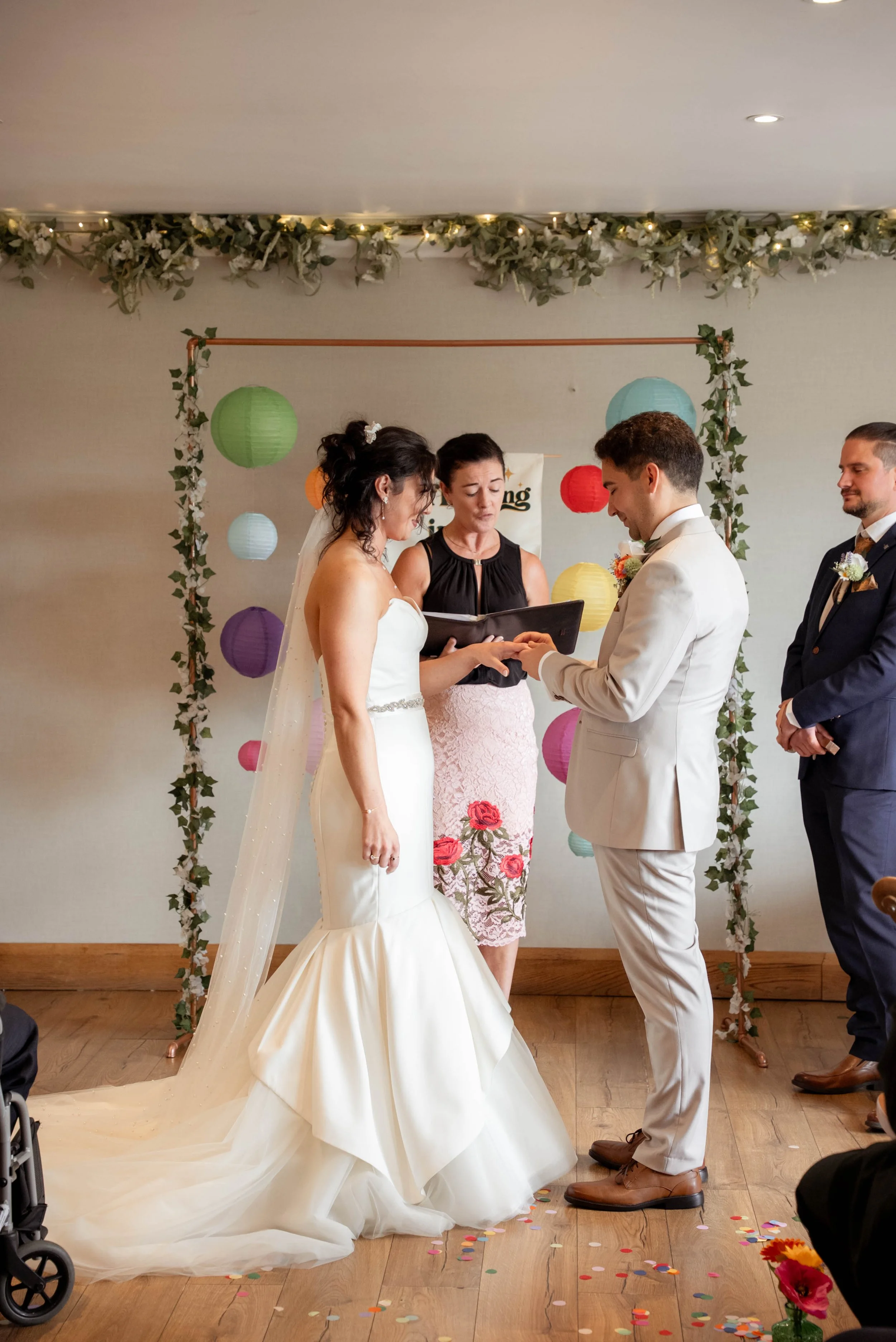 A bride and groom exchanging vows at their wedding ceremony, with an officiant and a groomsman present, decorated with colorful paper lanterns and greenery.