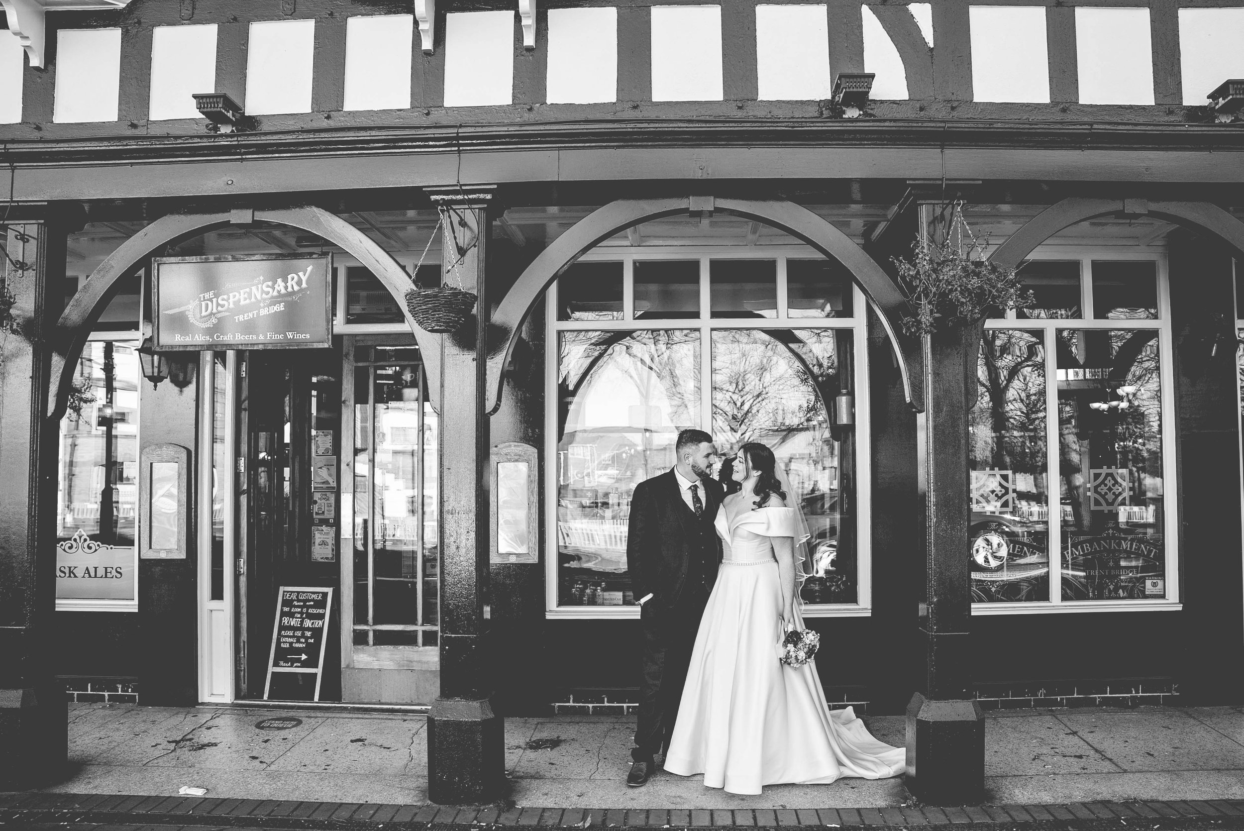 A black and white photo of a bride and groom standing outside a pub called The Dispensary, Trent Bridge. The bride is holding a bouquet and wearing a wedding gown, while the groom is in a suit. They are looking at each other affectionately.
