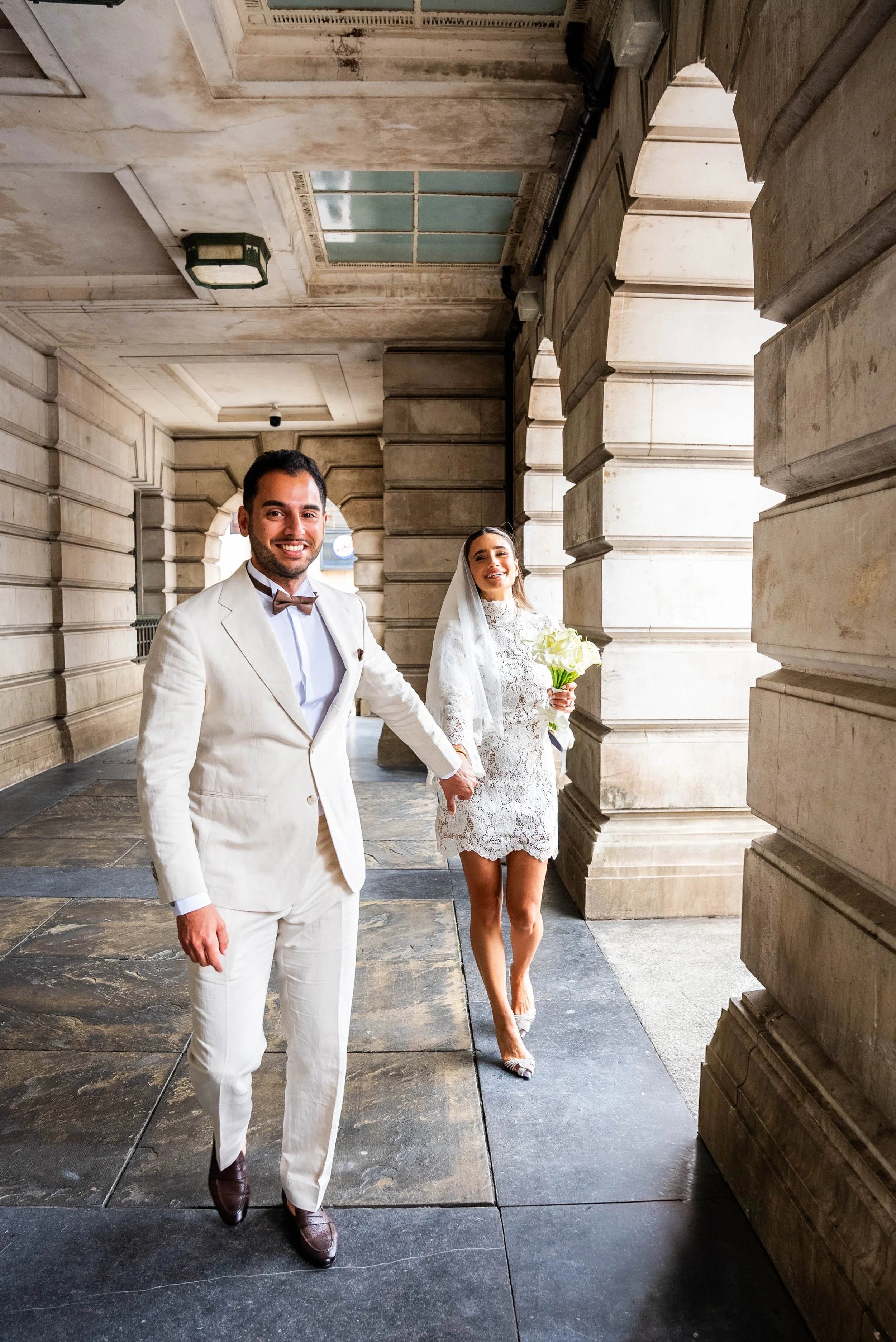 A happy newlywed couple walking hand in hand outdoors, with the bride holding a bouquet of white flowers, surrounded by stone columns and walls of a historic building.