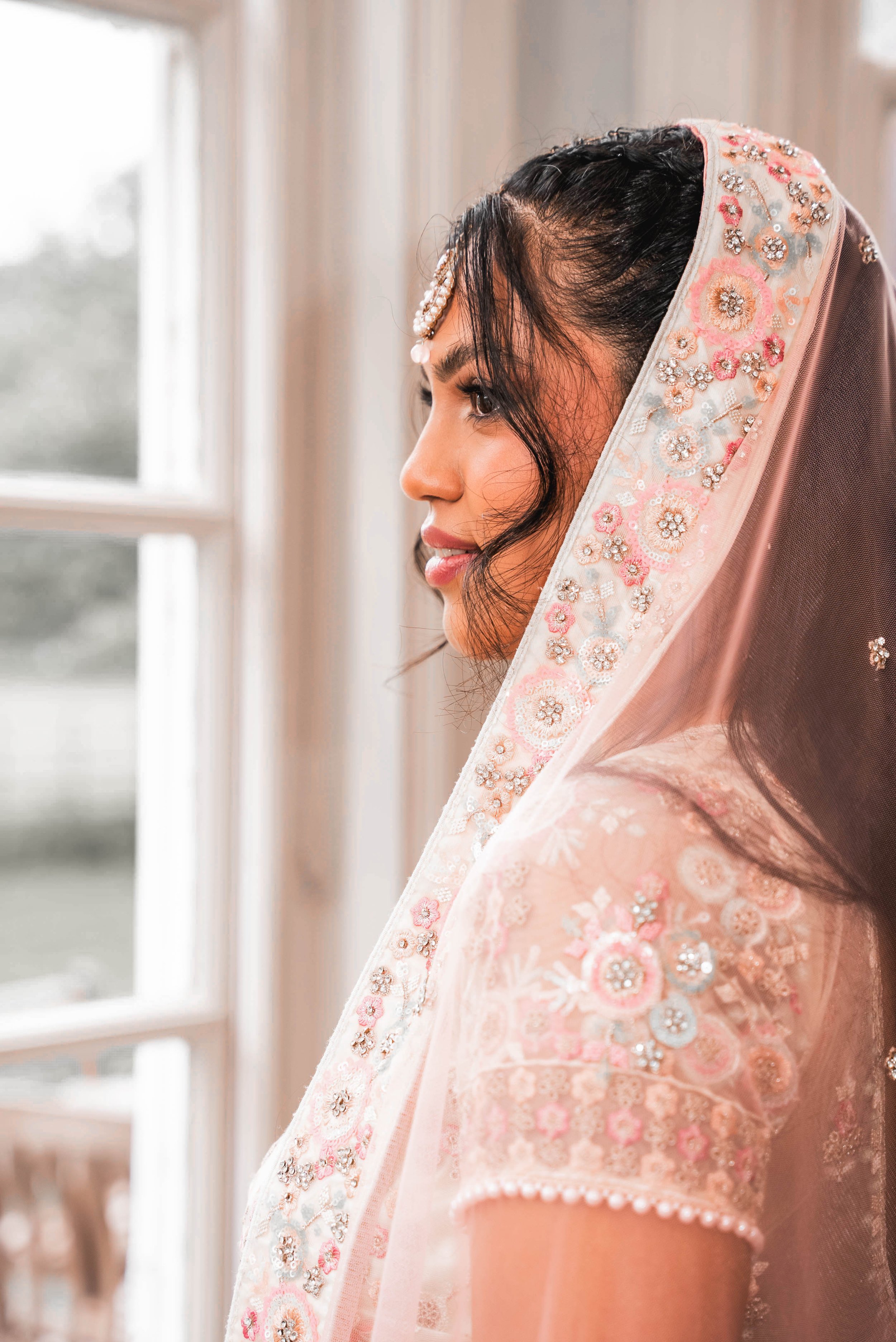 Close-up of a woman in traditional attire, wearing a pink, embroidered veil with floral and bead details, standing near a window with natural light.