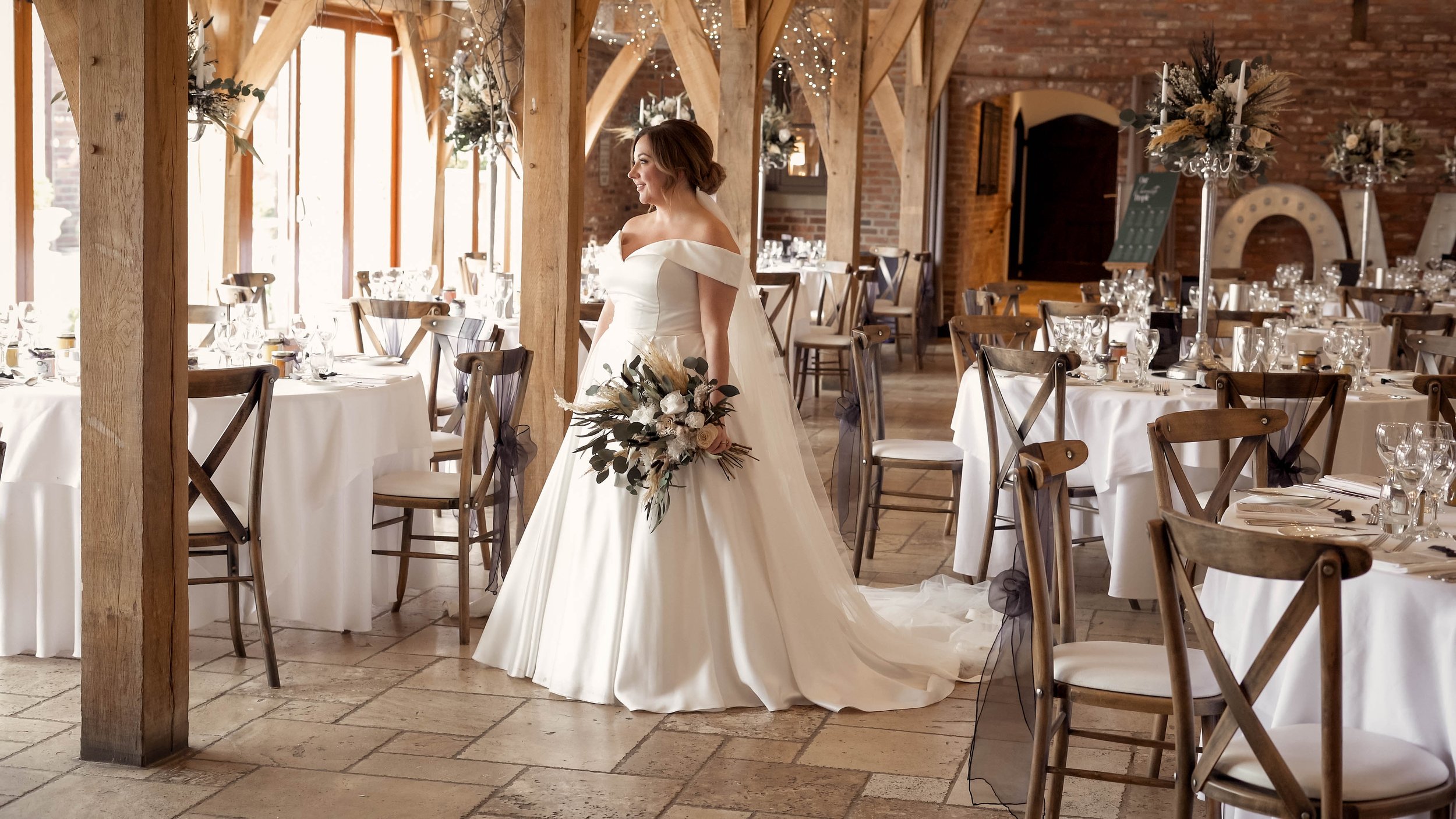 A bride in a white wedding gown holding a bouquet of flowers in a decorated banquet hall with round tables, white tablecloths, chairs, and floral centerpieces.