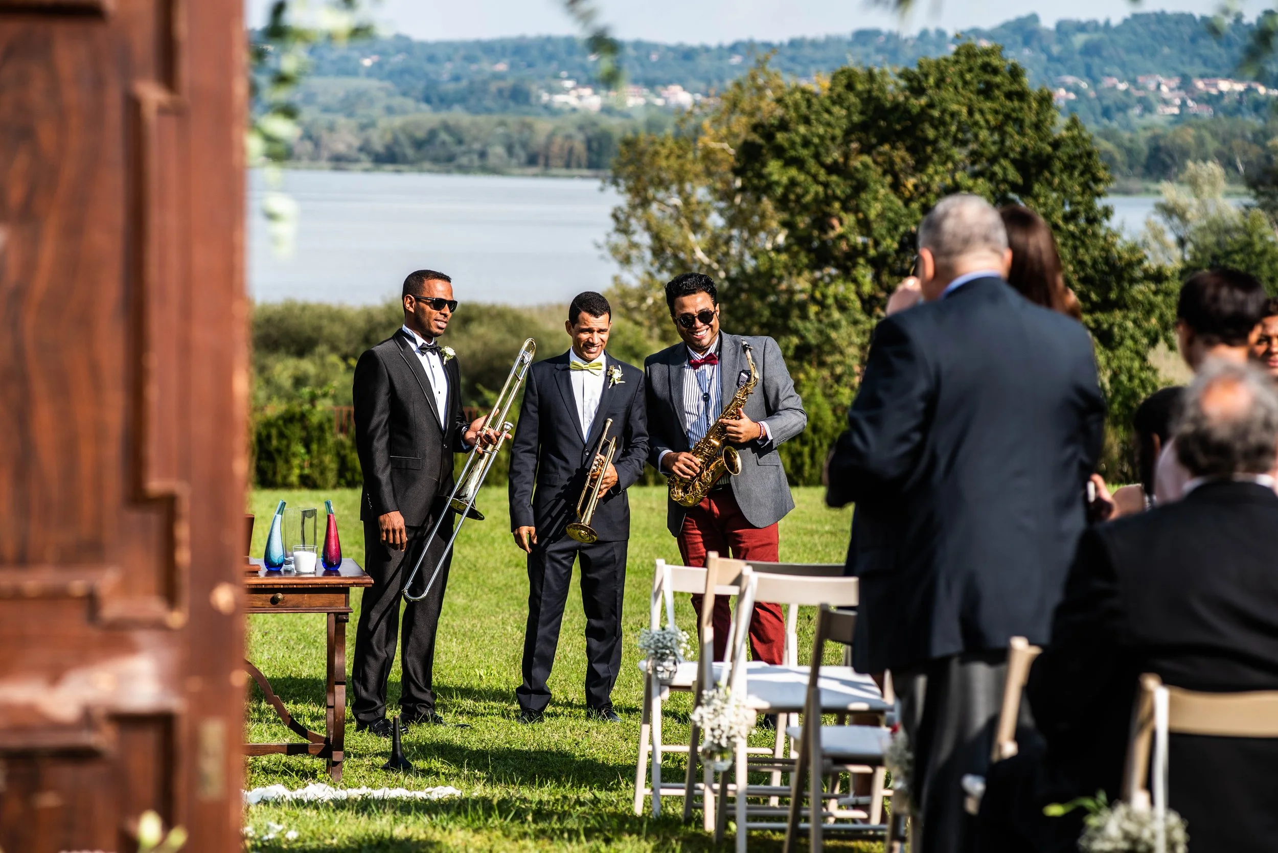 Musicians playing instruments at an outdoor wedding ceremony, with guests seated and overlooking a lake and green landscape