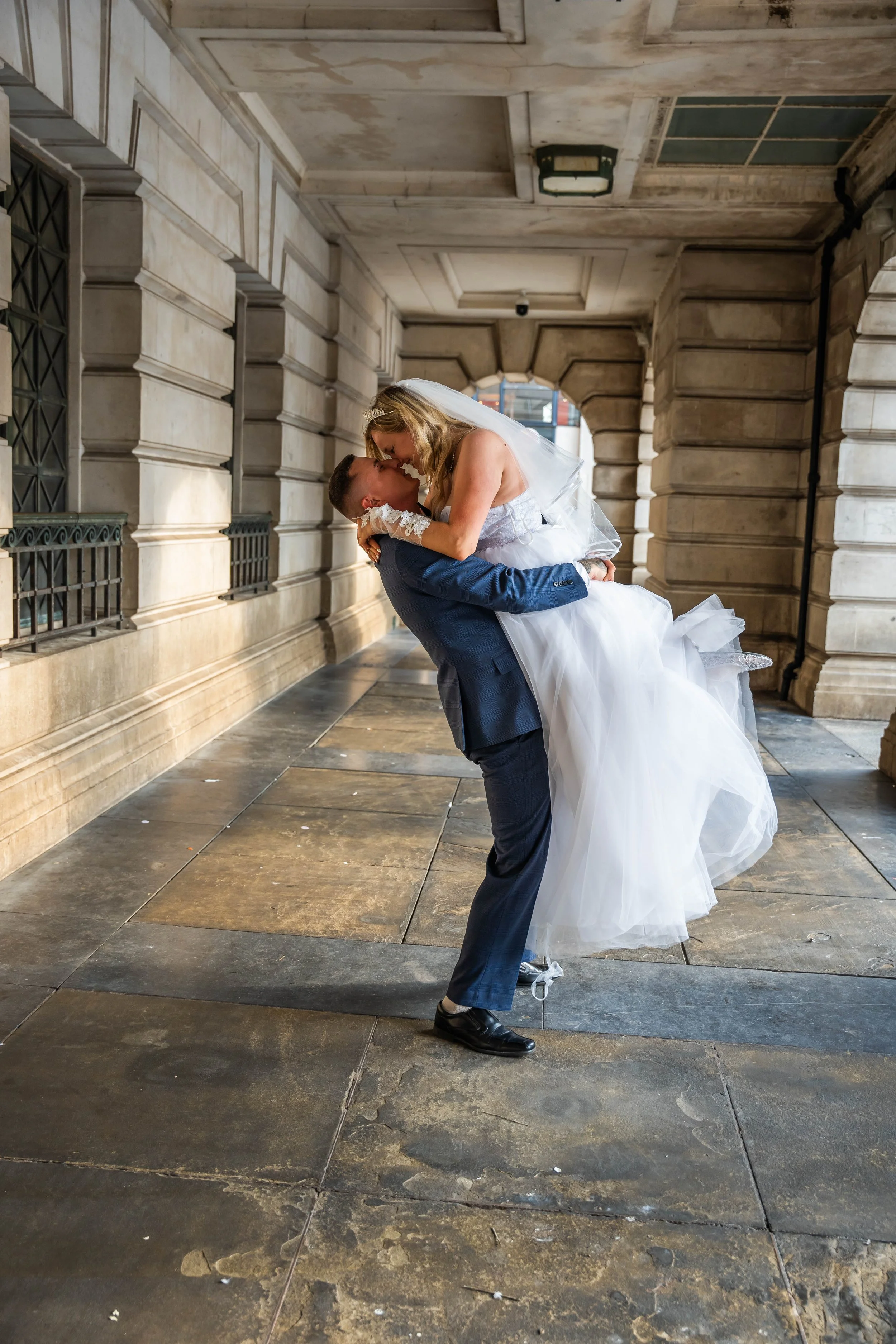 Groom carrying bride in a city sidewalk corridor, sharing a kiss, with stone walls and archways in the background.