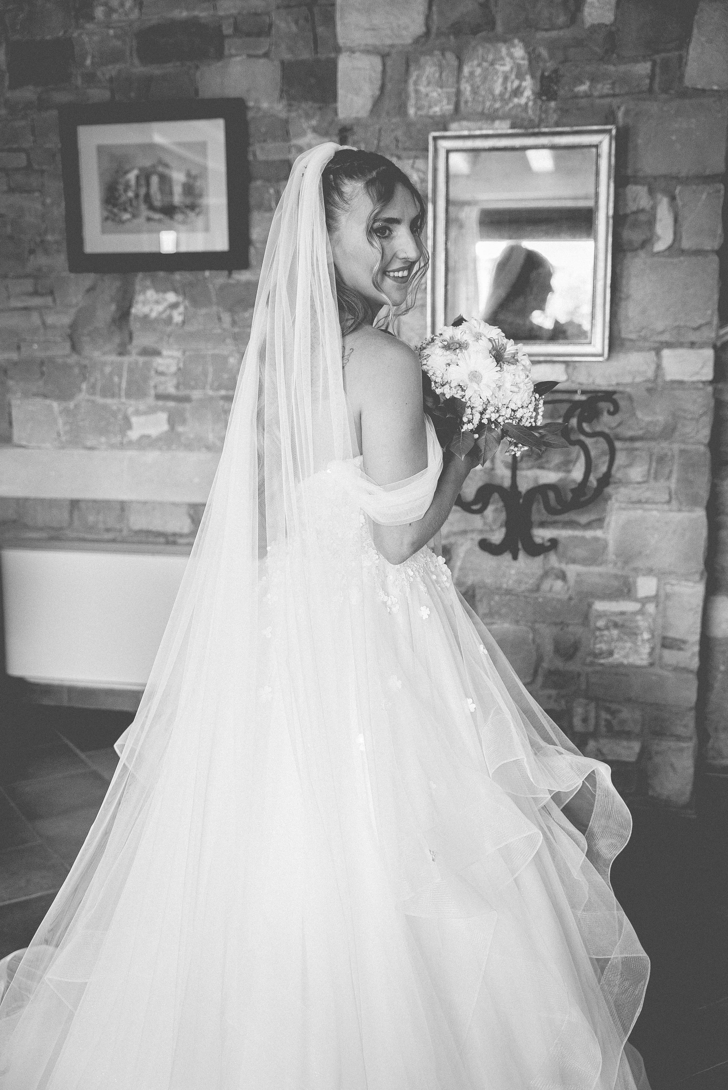 A bride in a wedding gown holding a bouquet of flowers, standing inside a room with a brick wall and framed pictures, smiling and looking over her shoulder.
