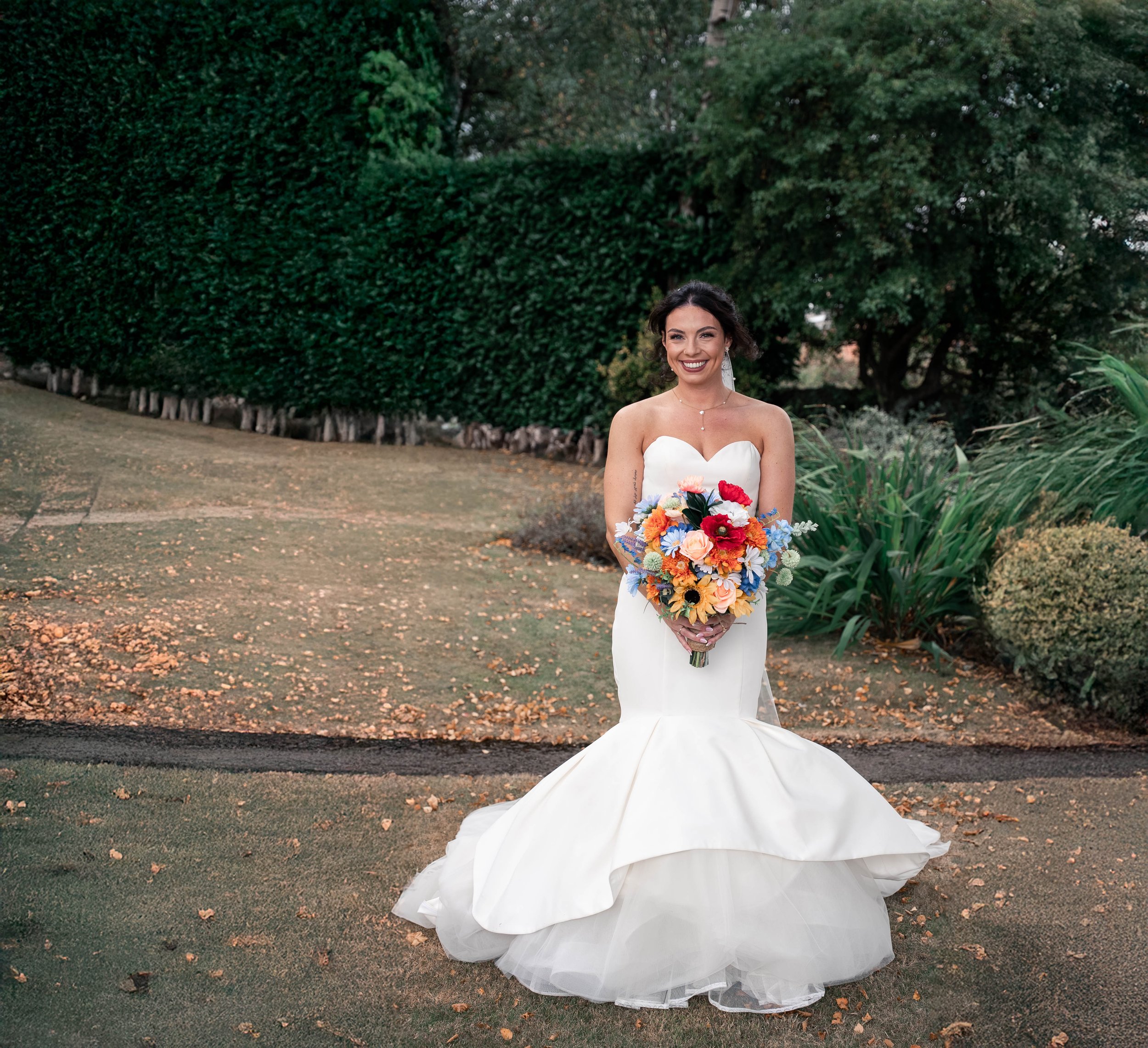 A smiling bride in a strapless white wedding gown with a flared skirt standing outdoors on a pathway, holding a vibrant bouquet of mixed colorful flowers, with lush green bushes and trees in the background.