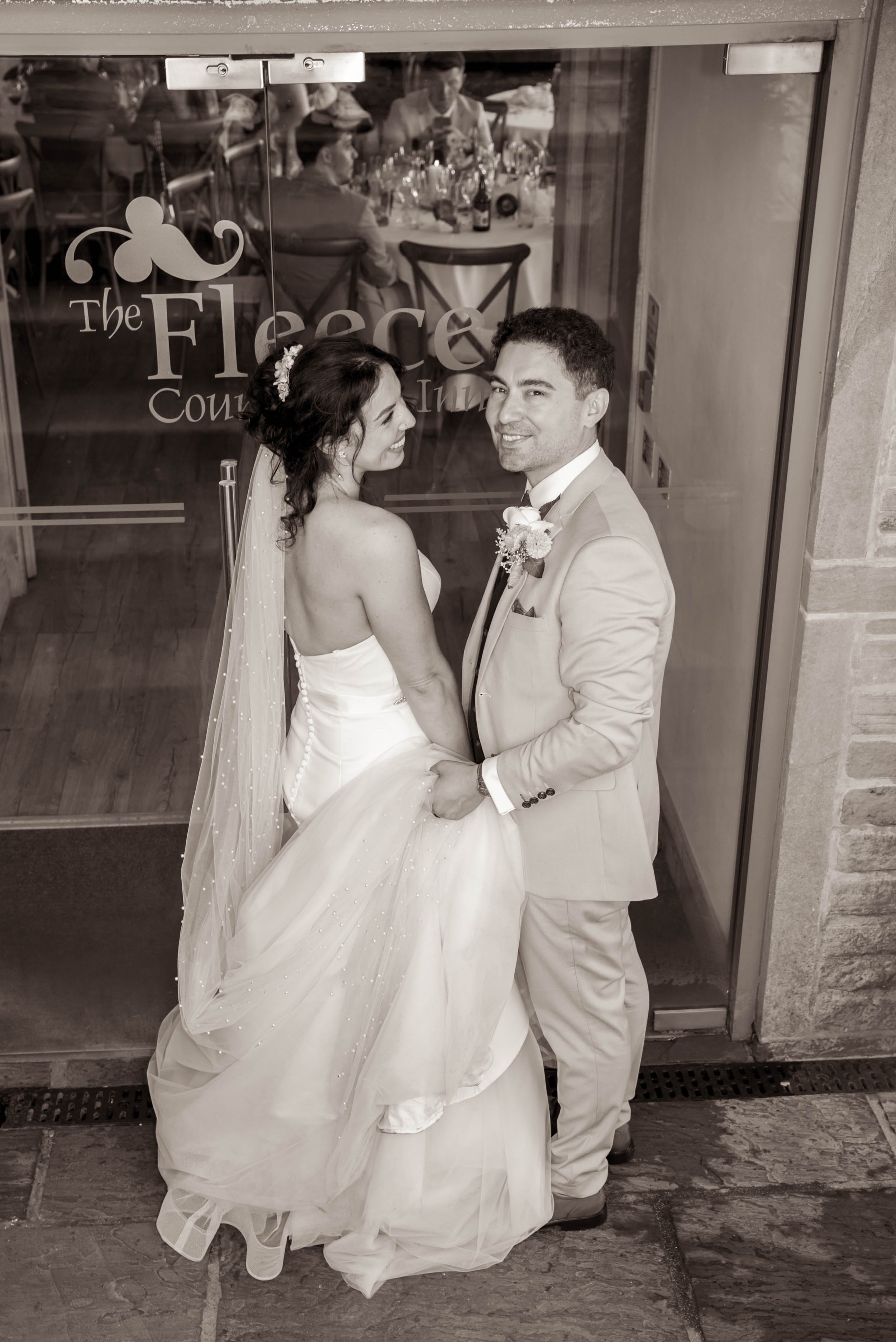 Black and white photo of a bride and groom standing outside a glass door, holding hands and smiling at the camera. The bride is in a strapless wedding dress with a long veil, and the groom is in a light-colored suit with a boutonniere. Reflected gues