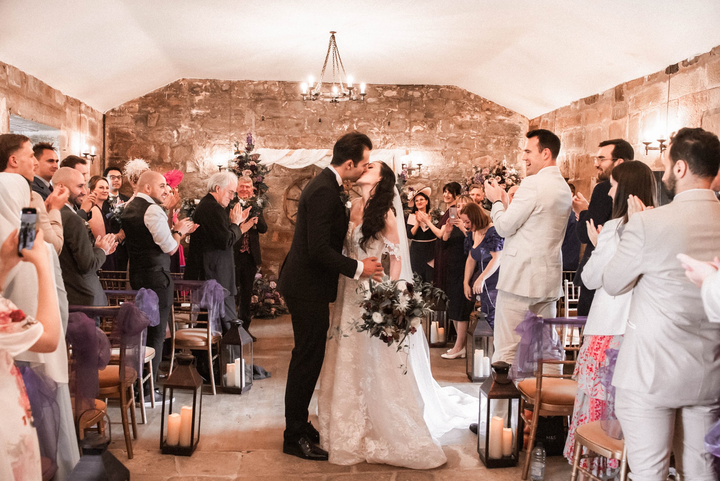 A wedding ceremony with a bride and groom kissing in front of guests inside a rustic stone-walled venue, decorated with floral arrangements and candles.