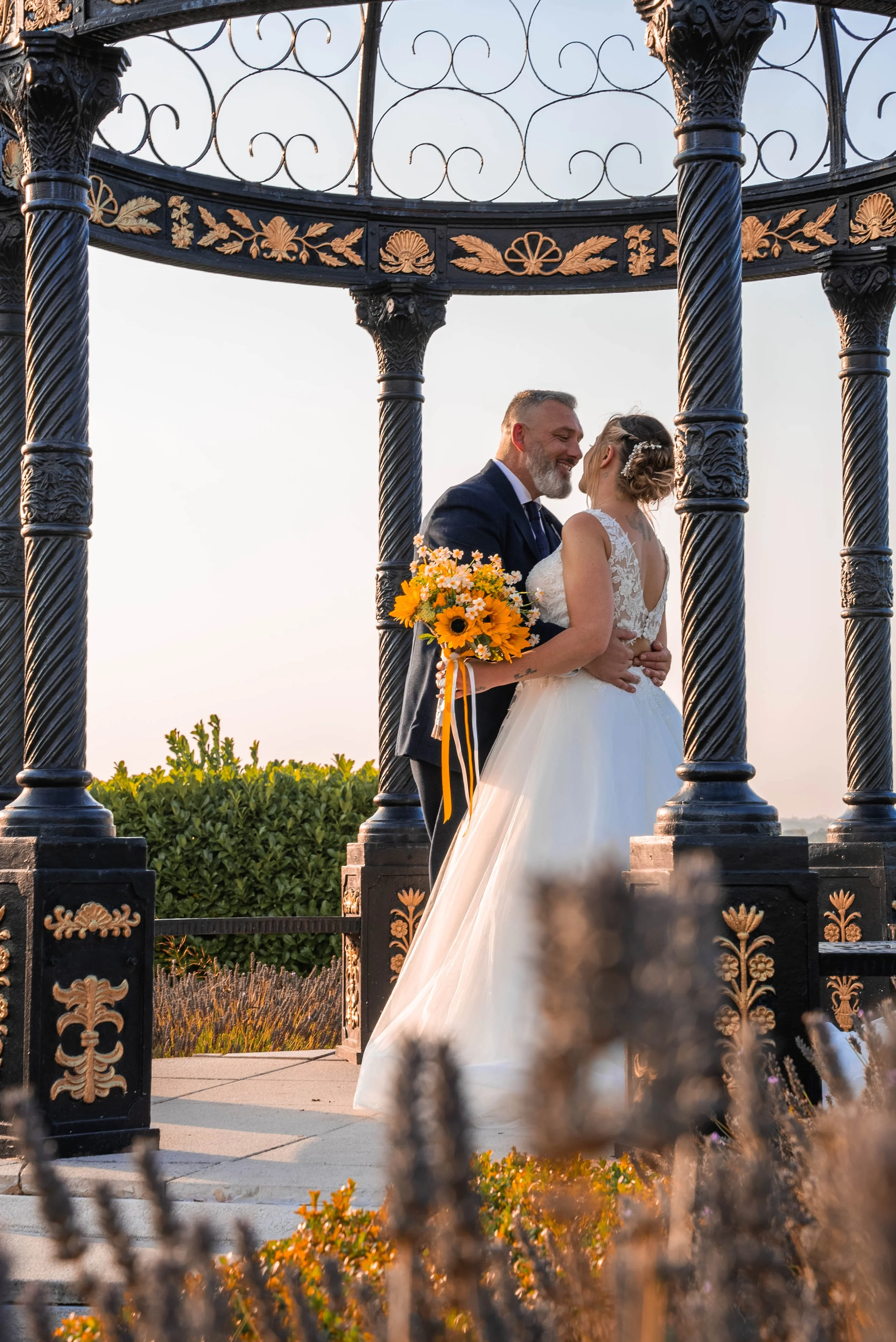 A bride and groom share a romantic moment under an ornate black and gold gazebo during their wedding, with the bride holding a bouquet of yellow flowers and wearing a white lace wedding gown with a tulle skirt. The groom is dressed in a dark suit, an