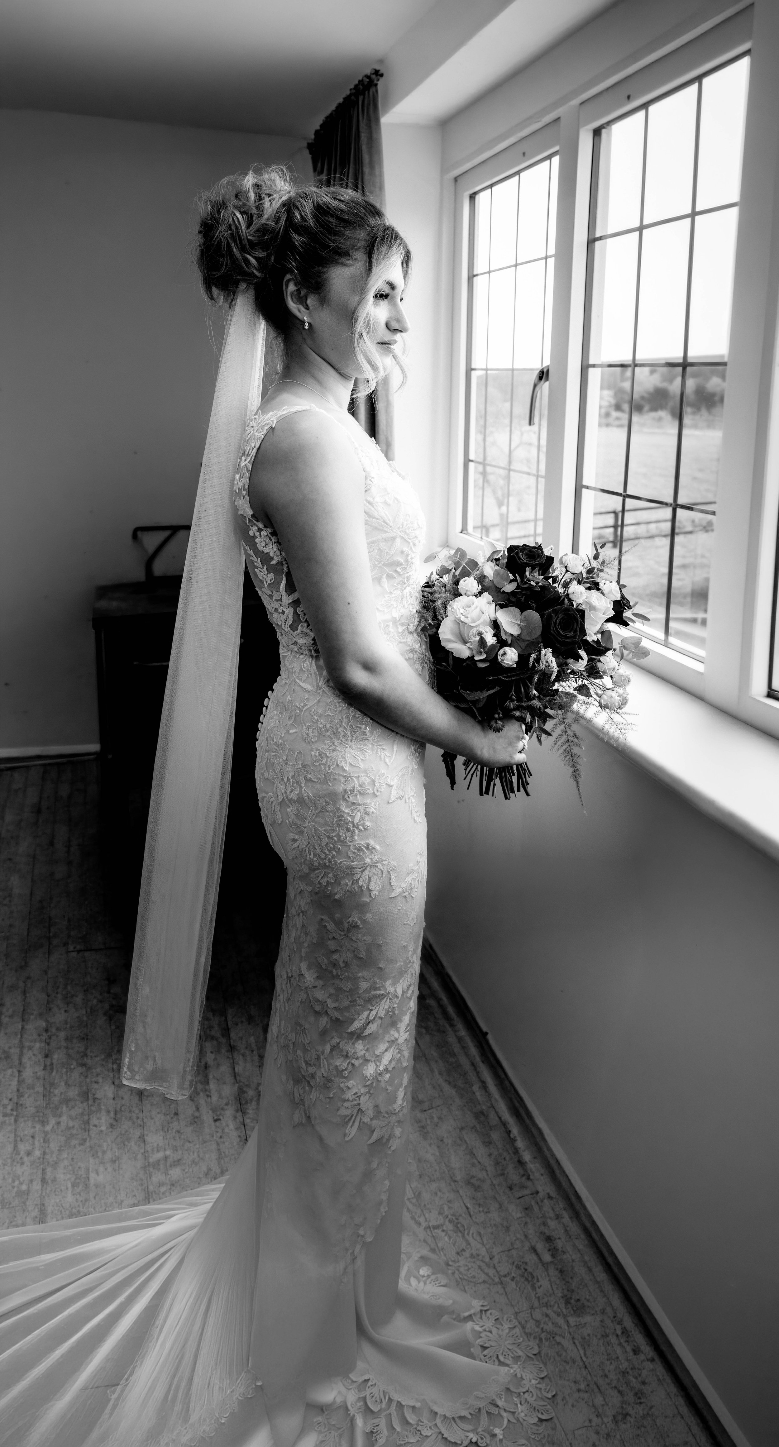 A bride in a lace wedding gown holding a bouquet of dark and light flowers, standing by a window with a view of a field or yard outside, in a room with wooden floor and curtains.