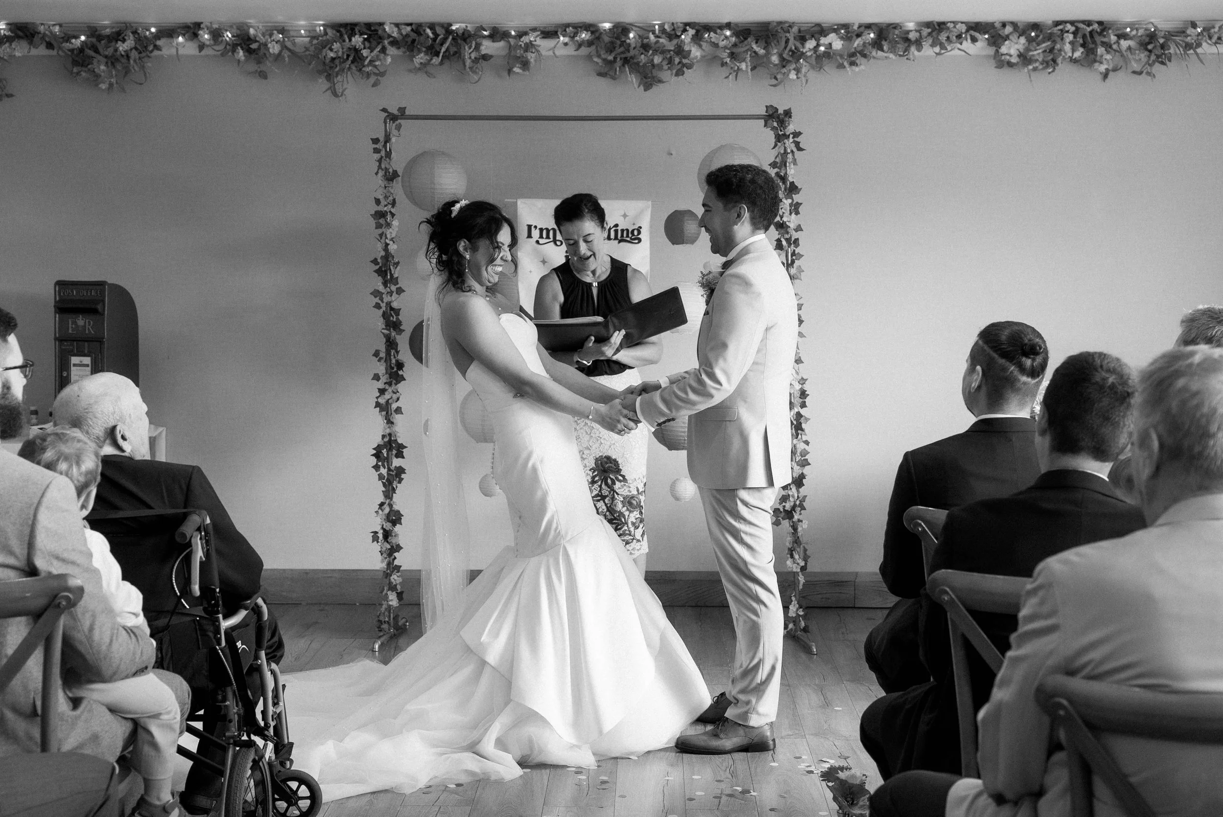 A black-and-white photo of a wedding ceremony with a bride and groom holding hands, standing before an officiant. The bride wears a wedding gown with a veil, and the groom is in a light suit. Guests are seated on either side, watching the ceremony in