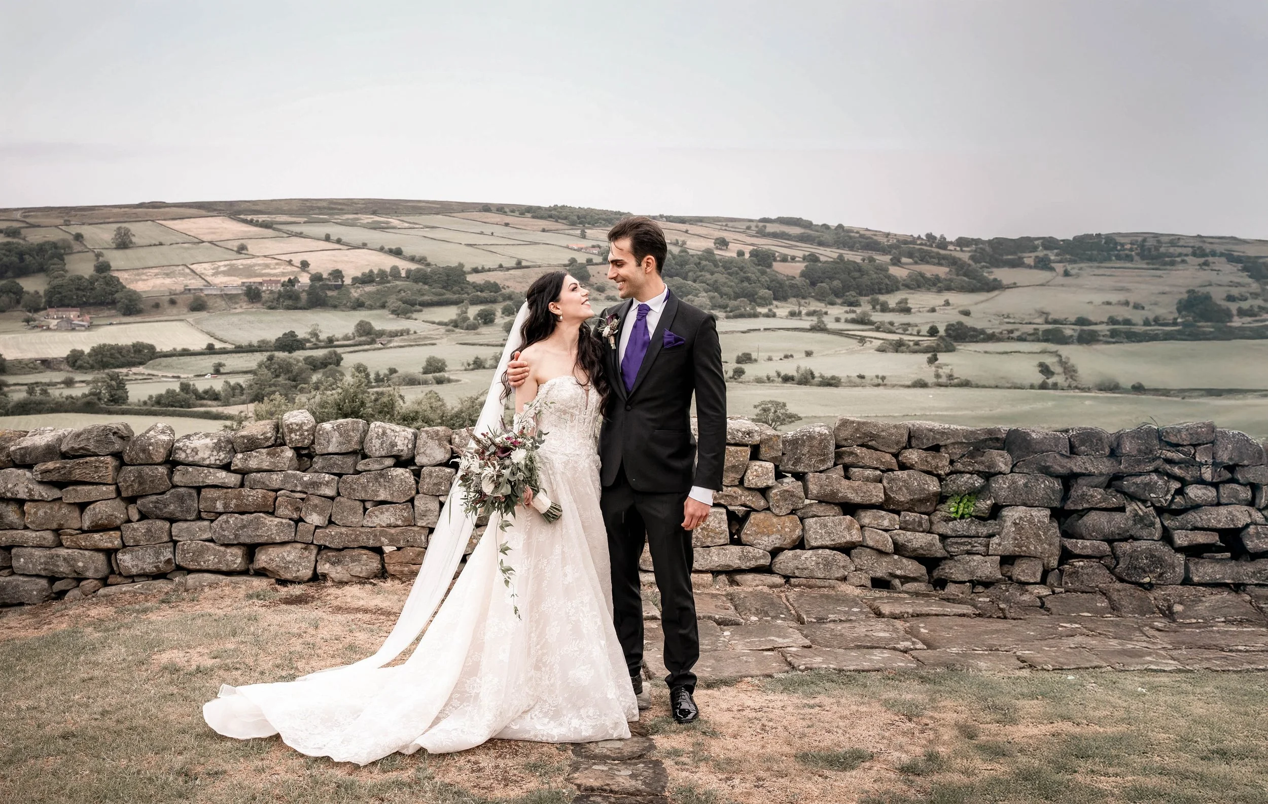 Wedding couple standing together outdoors with stone wall and rolling green hills in the background.