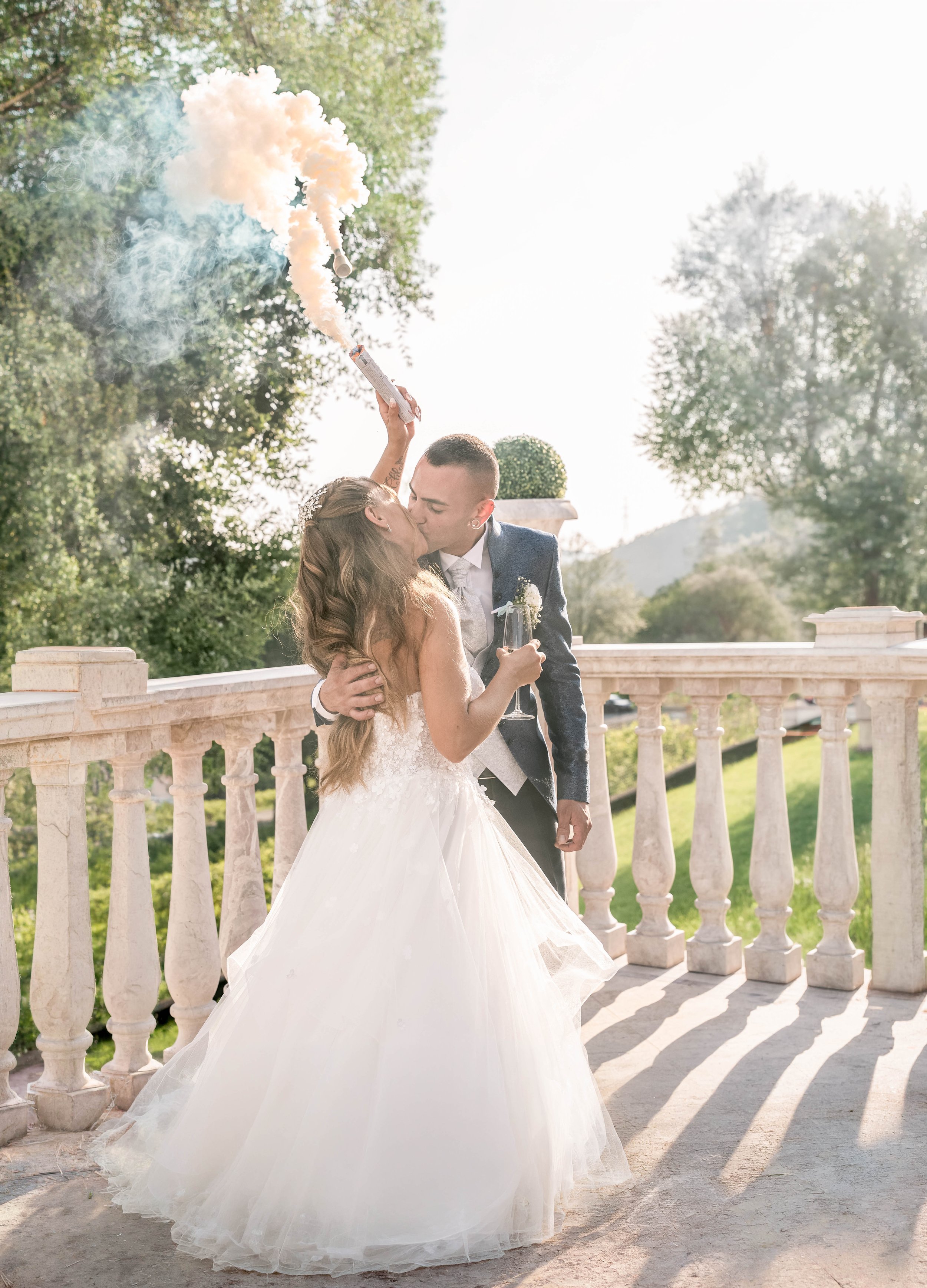 A bride and groom sharing a kiss on a balcony with balustrades, surrounded by trees and natural scenery during the daytime. The bride is wearing a white wedding dress and the groom is in a suit.