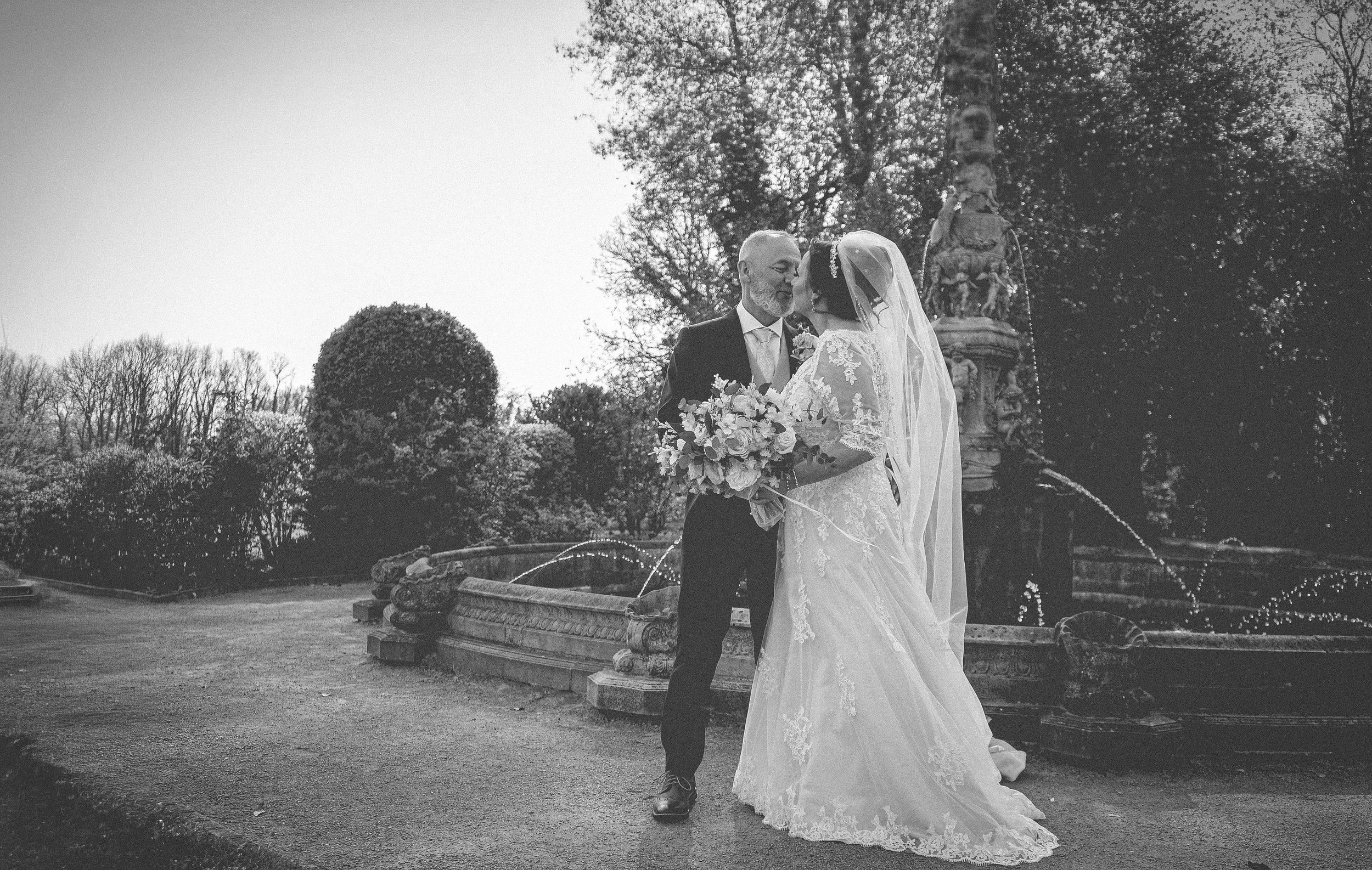 A bride and groom stand close to each other in front of a decorative fountain, sharing a moment on their wedding day. The bride holds a bouquet and wears a lace wedding gown with a long veil, while the groom is dressed suit-style.
