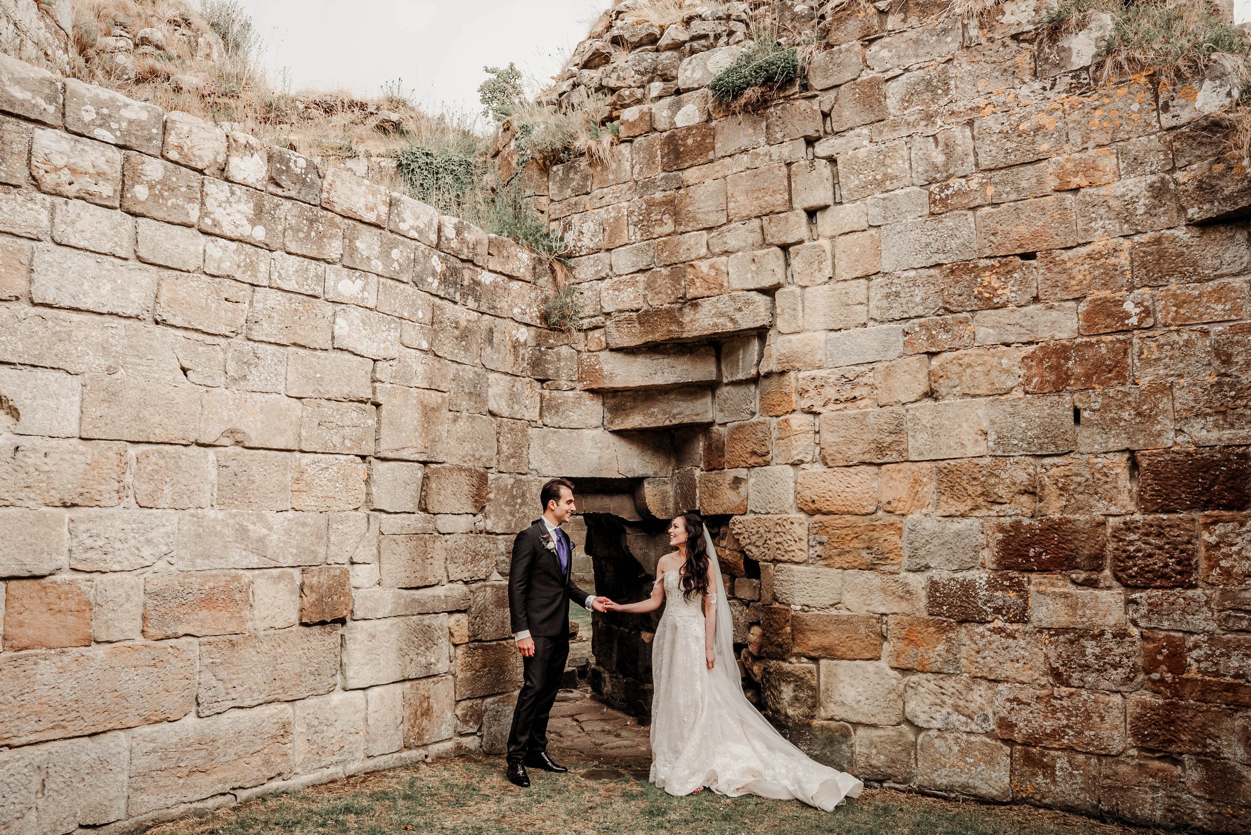 A bride and groom in wedding attire holding hands and smiling at each other, standing inside ancient stone walls on a grassy area.