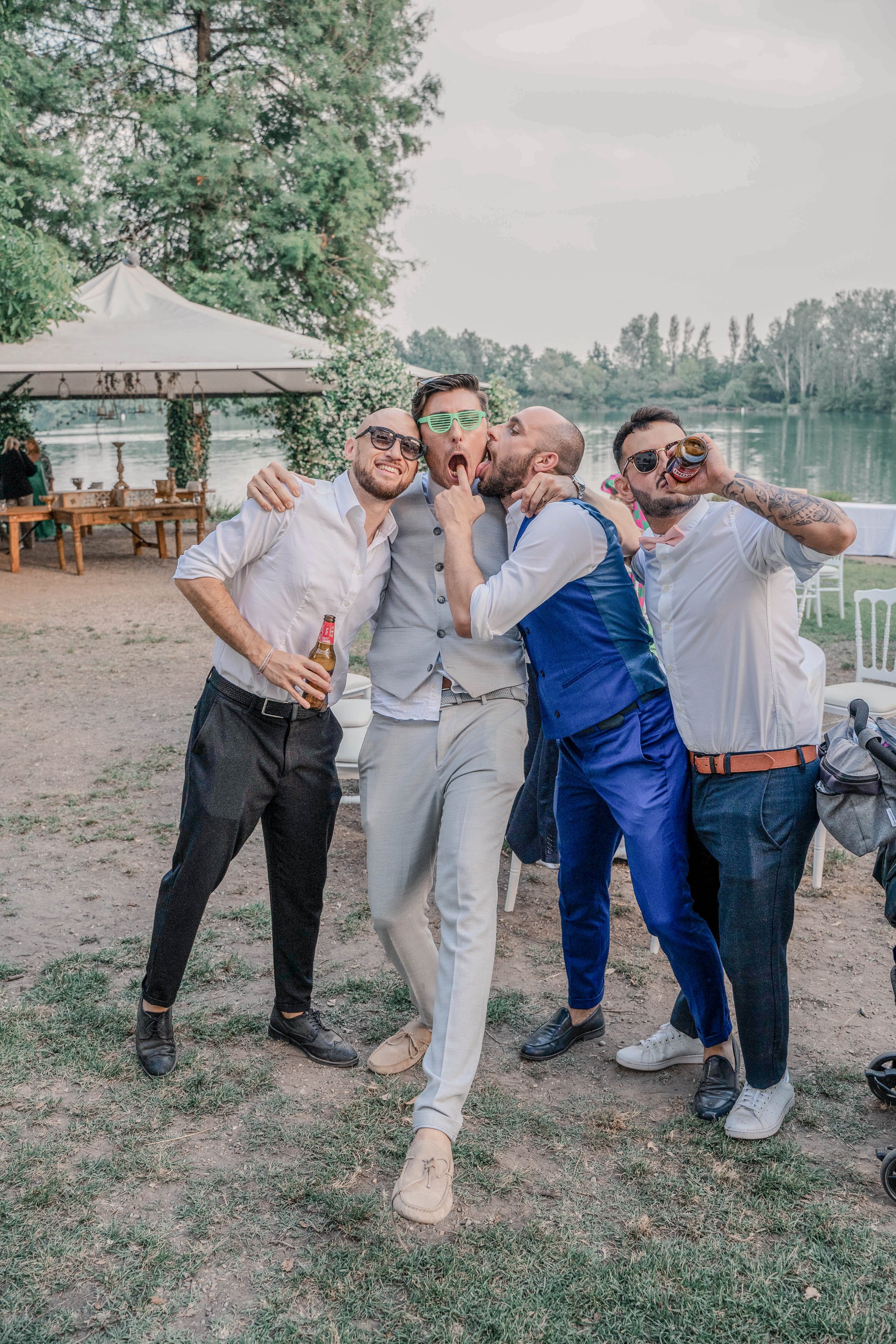 Group of five men enjoying an outdoor social event by a river, with some holding drinks and making playful gestures.