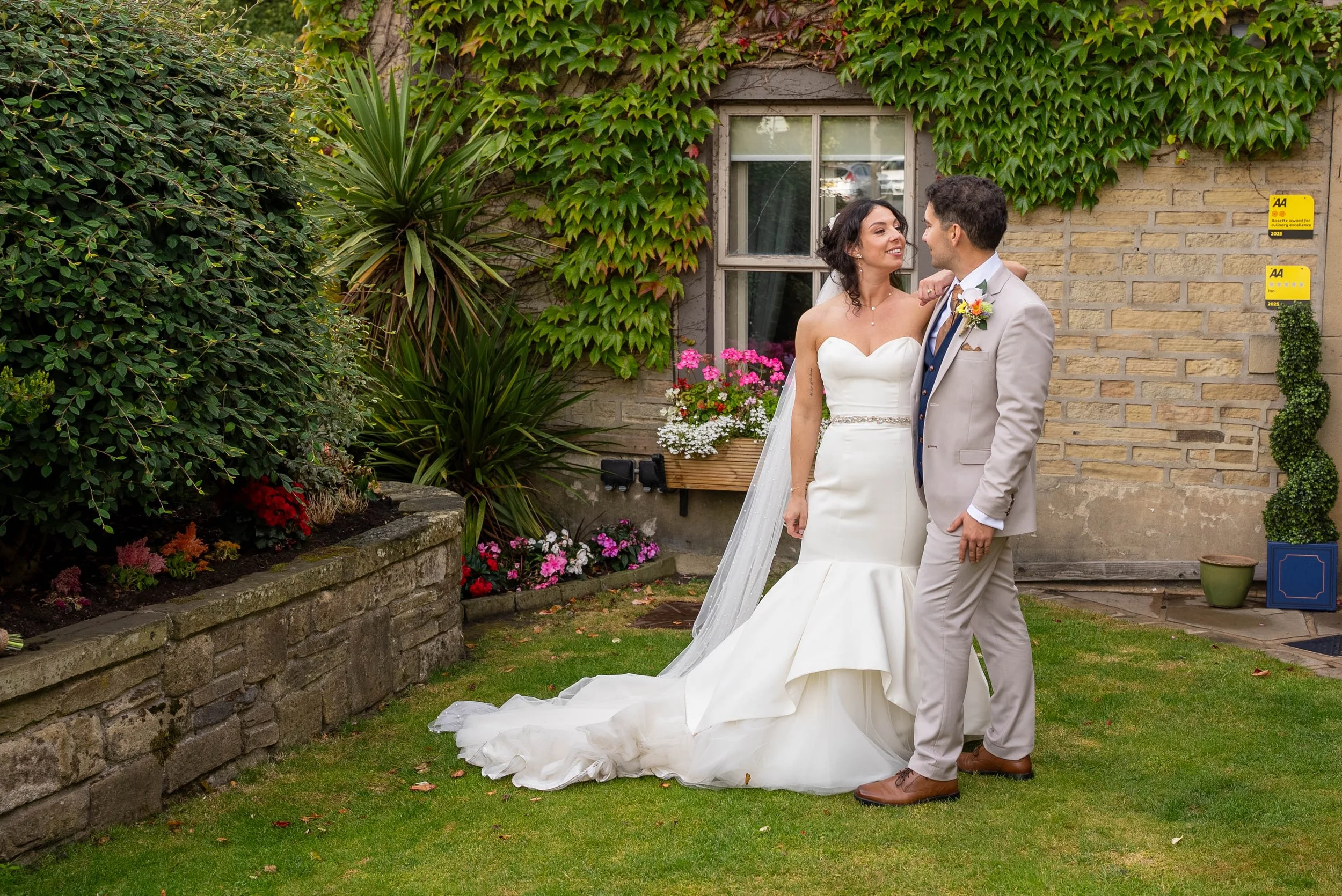A bride and groom standing together in a garden, looking at each other. The bride is wearing a strapless white wedding gown with a train, and the groom is wearing a light-colored suit with a blue tie. There are colorful flowers and green foliage behi