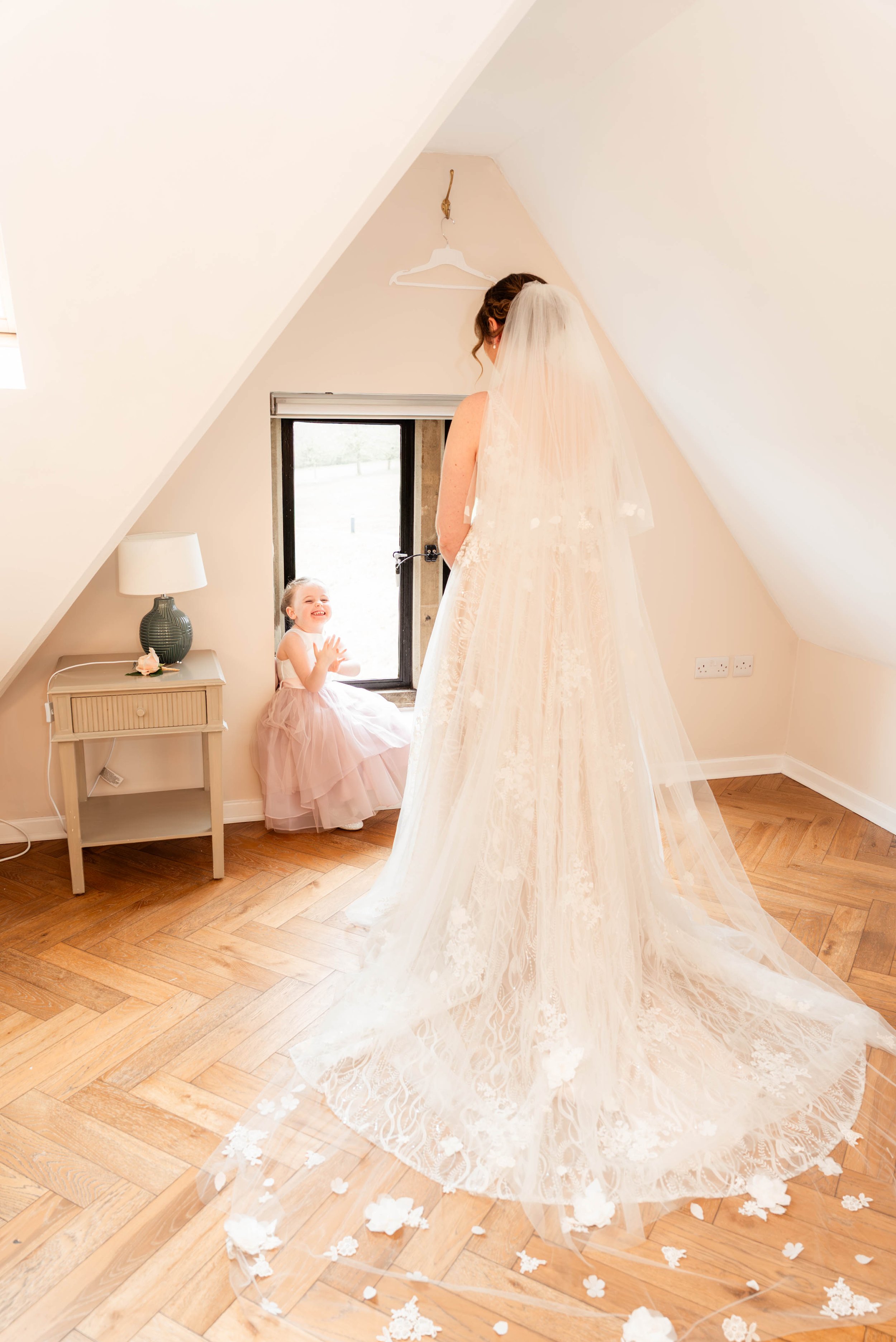 A bride in a wedding dress with a long veil standing in a room with wooden floors, facing a young girl sitting on a window ledge, who is smiling and clapping. The room has sloped ceilings and a small bedside table with a lamp and a flower on it.