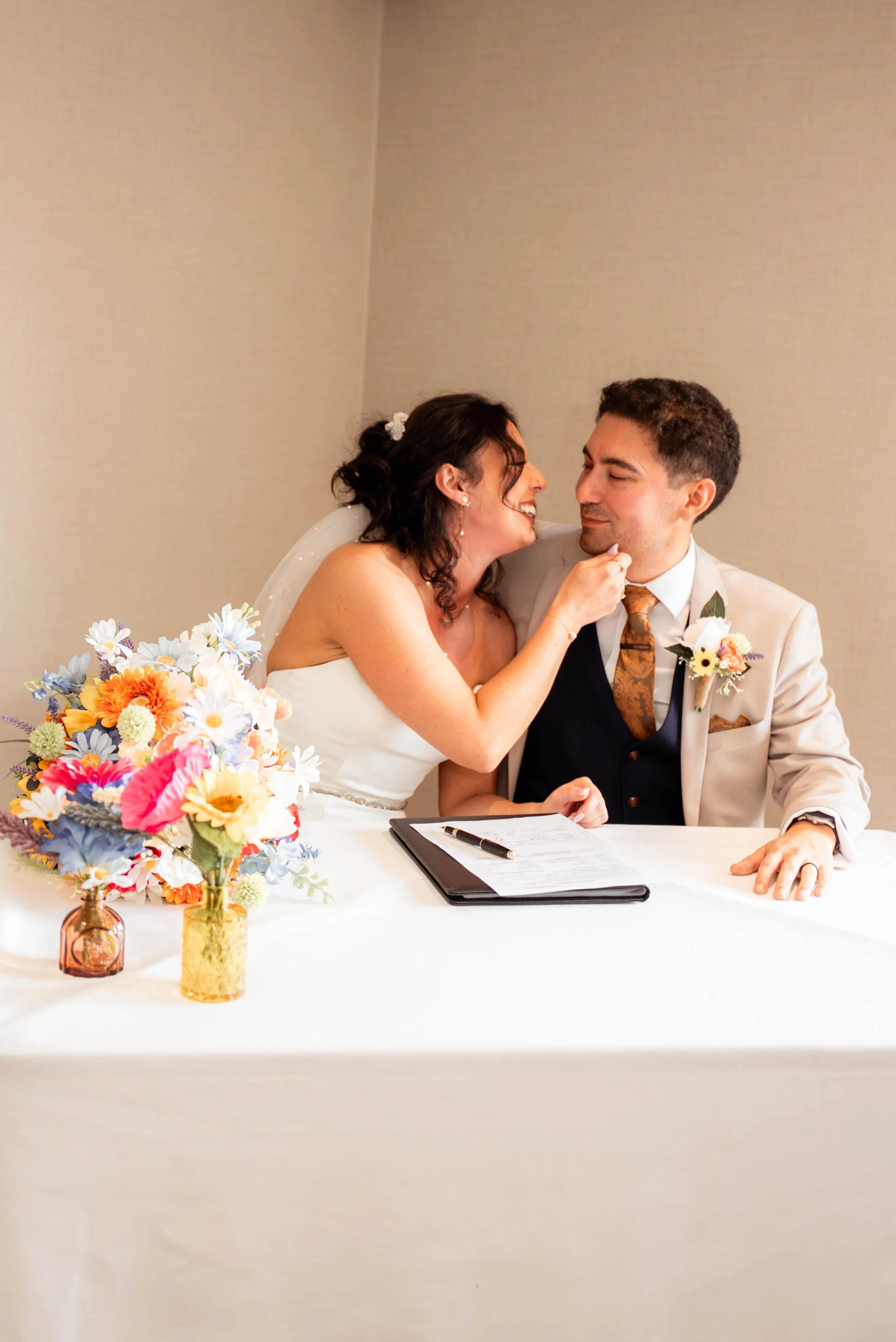 A smiling bride and groom sit at a table during their wedding ceremony, with the bride gently touching the groom's chin. There is a colorful bouquet of flowers on the table beside them.