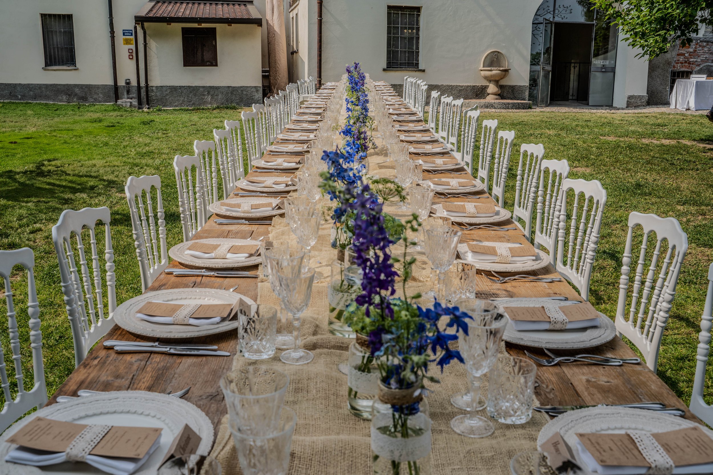 Long outdoor banquet table set for a meal, decorated with blue flowers in vases, with white chairs and table settings, situated on a grassy yard in front of a building.