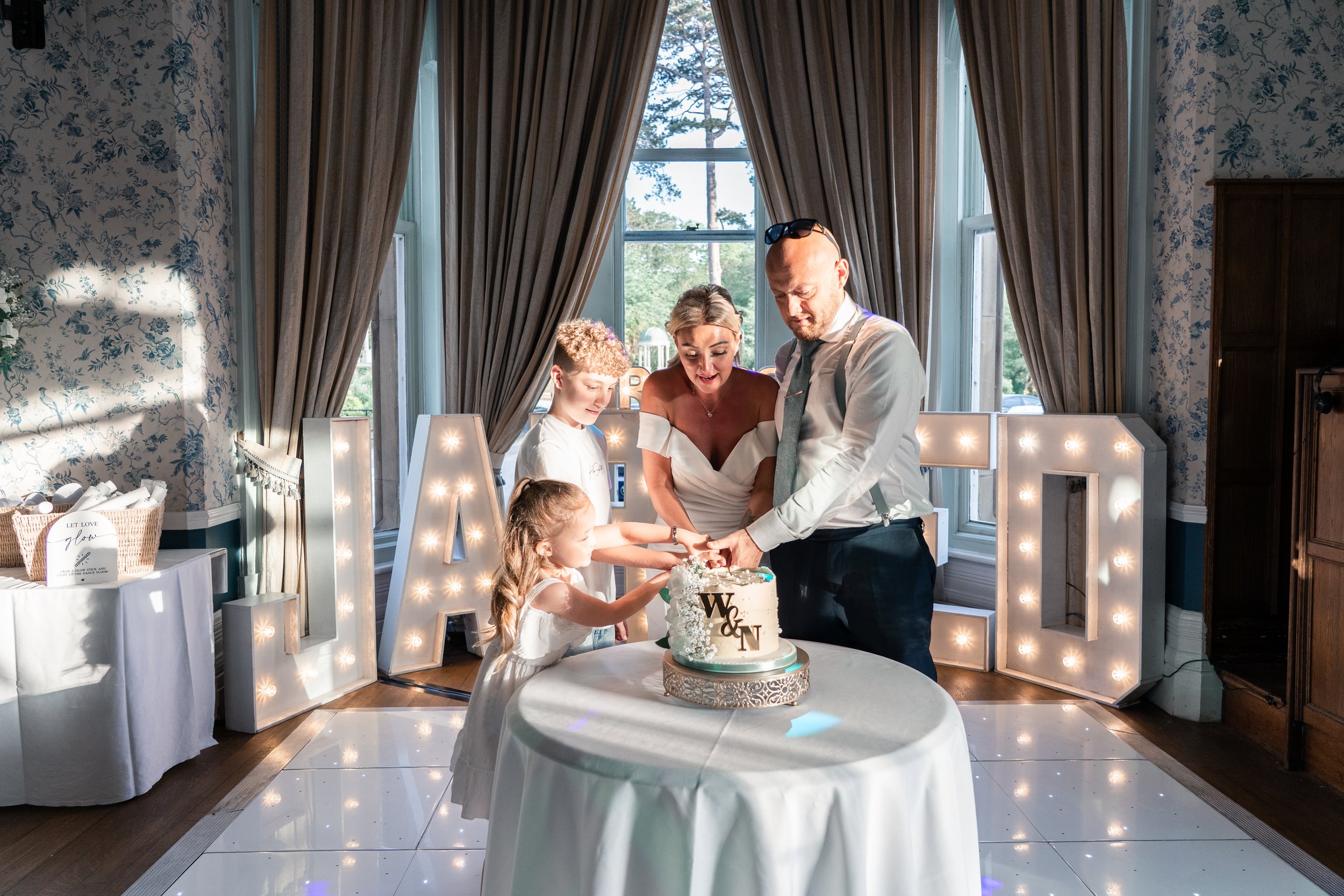 A family celebrating a birthday with a cake in a decorated indoor space with large windows, curtains, and illuminated letters in the background.