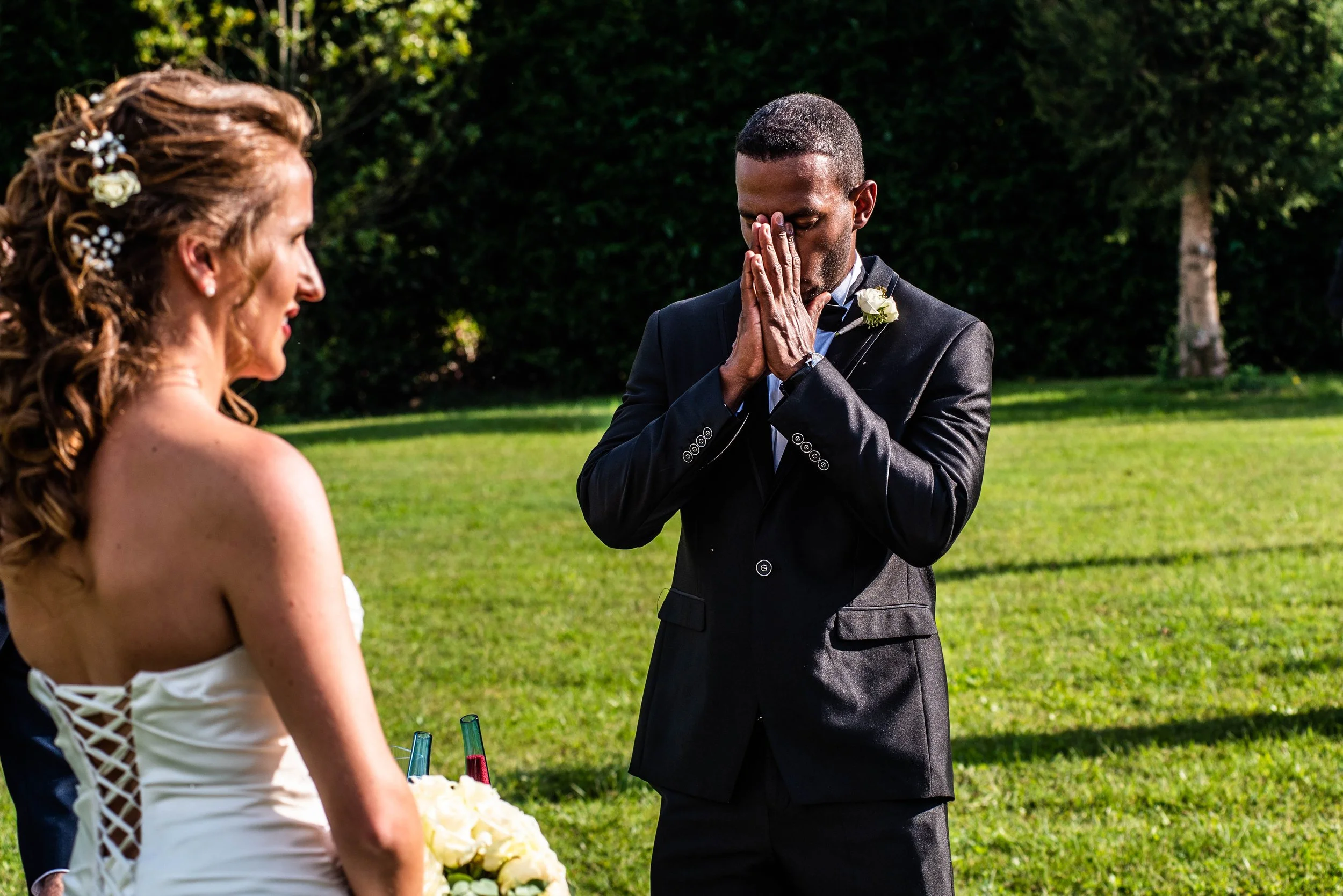 A bride and groom exchange vows outdoors during a wedding ceremony. The groom appears emotional, with hands clasped near his face, while the bride looks on.