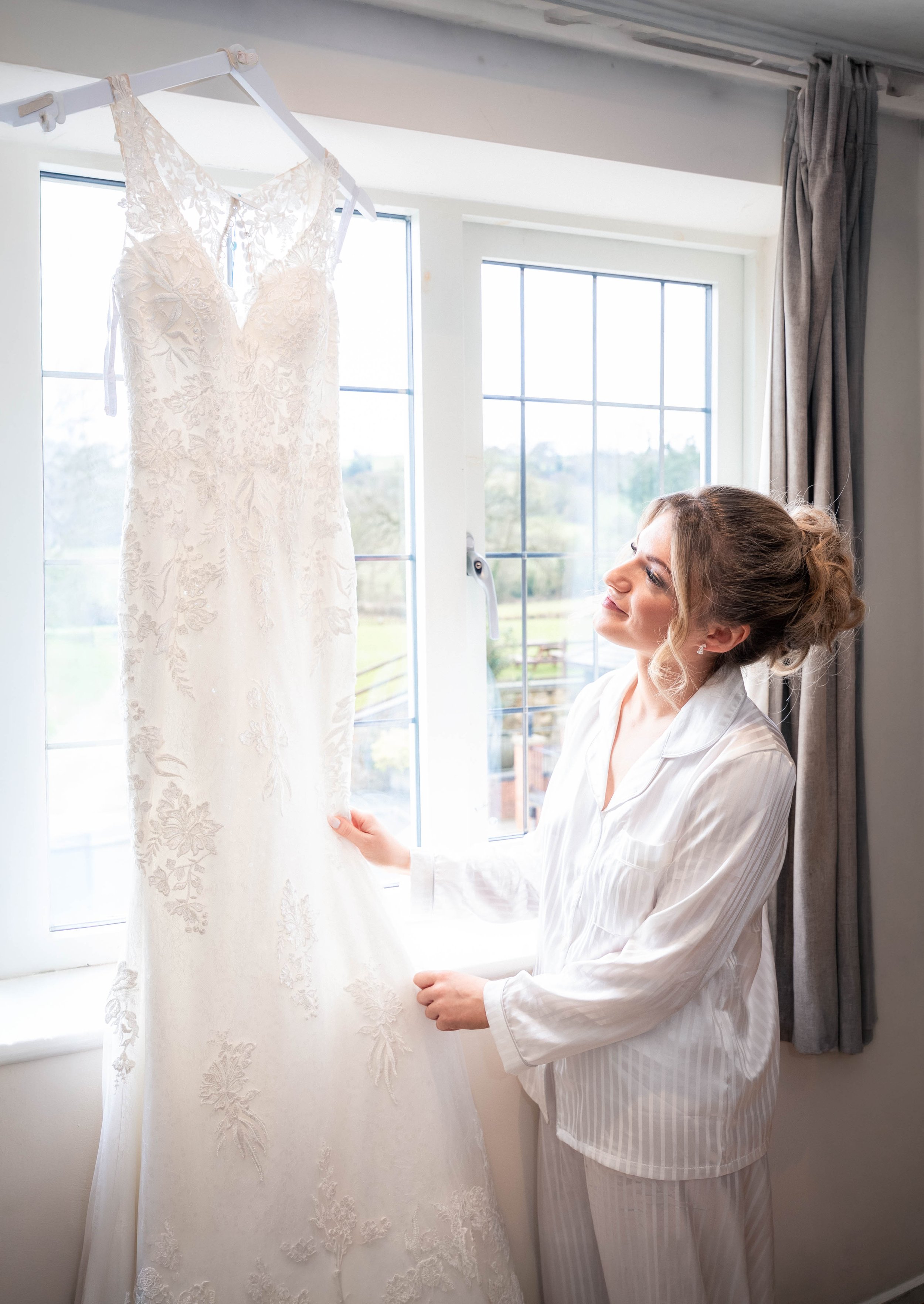 Bridal bride in pajamas holding her wedding dress in front of a window.