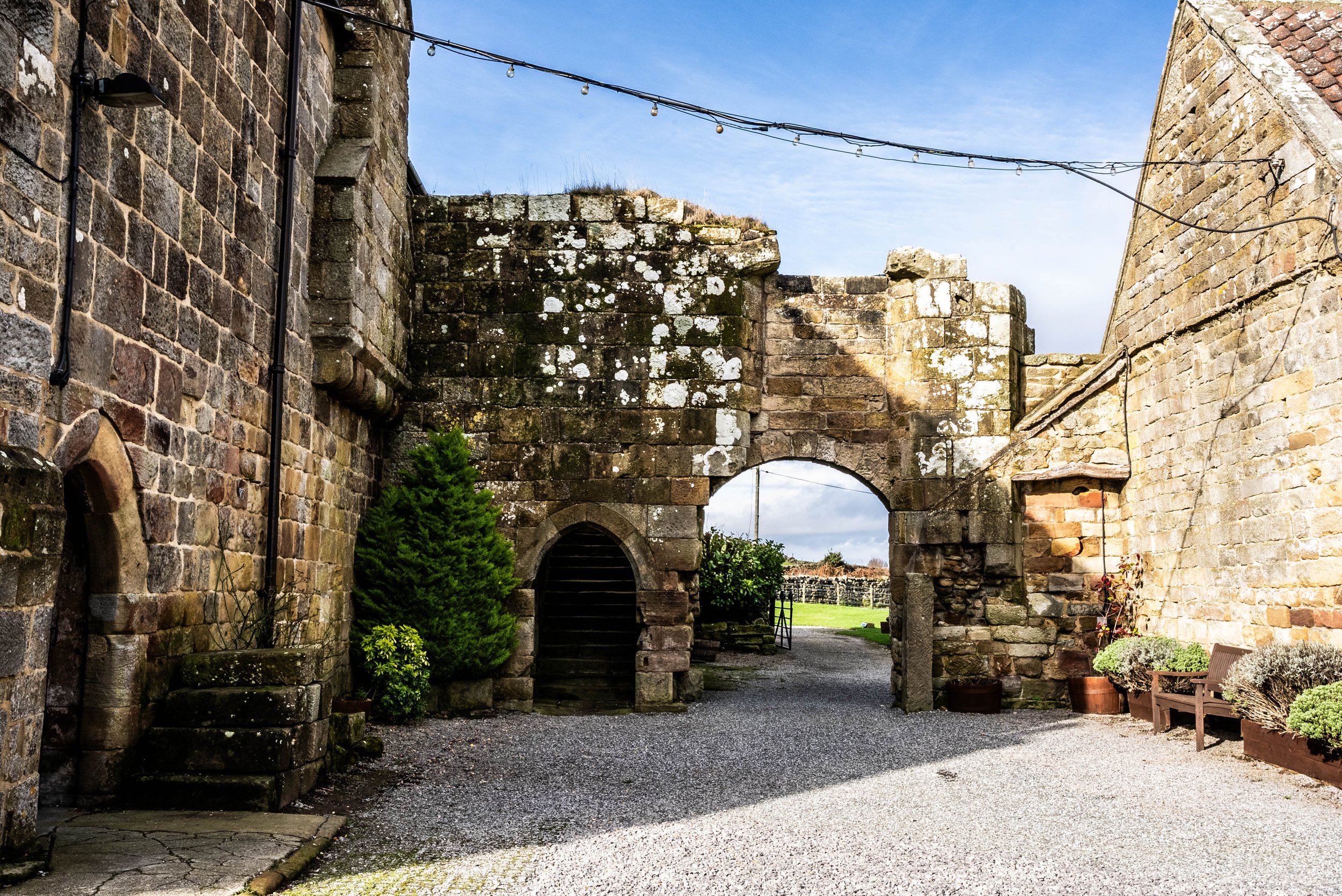An architectural scene featuring old stone walls and an archway with stairs leading up through it, a small garden with potted plants, a bench, and string lights hanging above on a bright day.