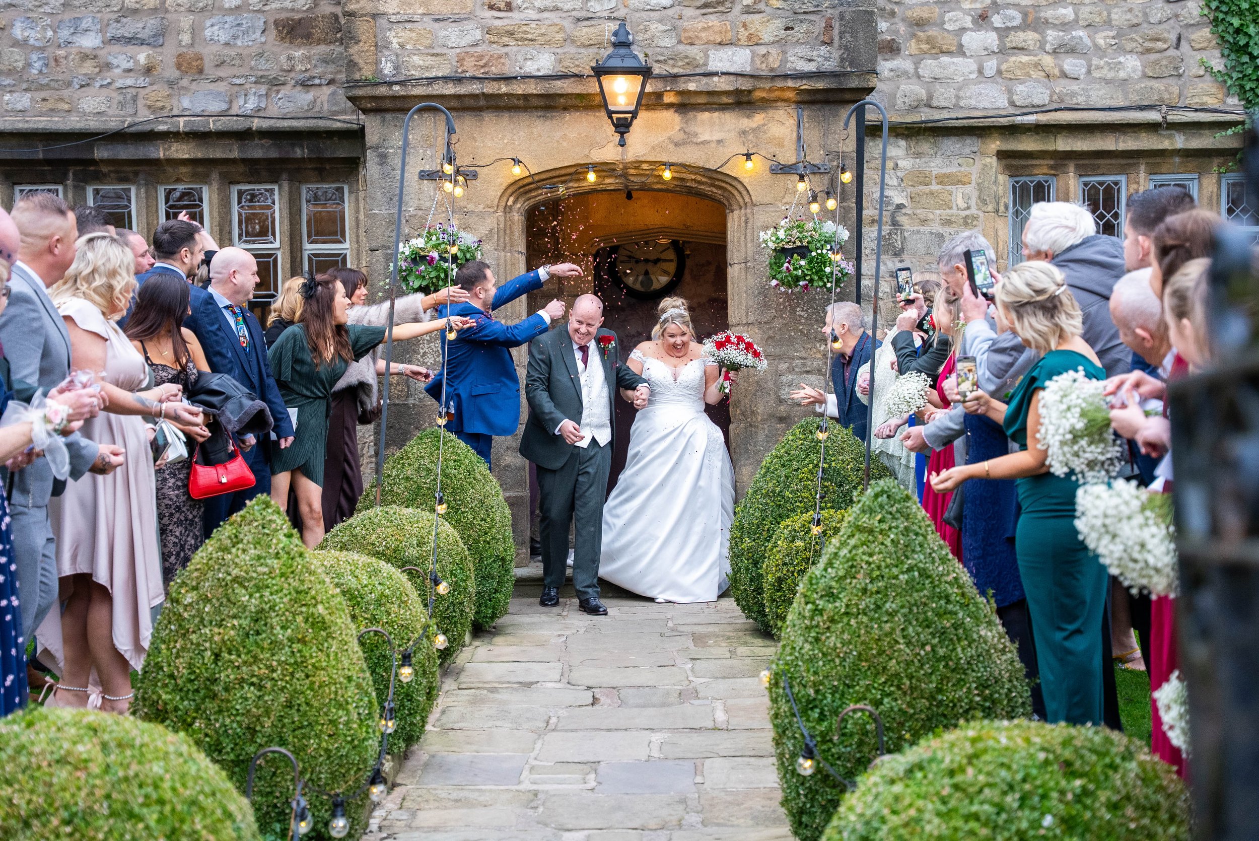 A bride and groom exit a church while surrounded by guests, with the bride holding a bouquet and the groom in a gray suit, as confetti is thrown.