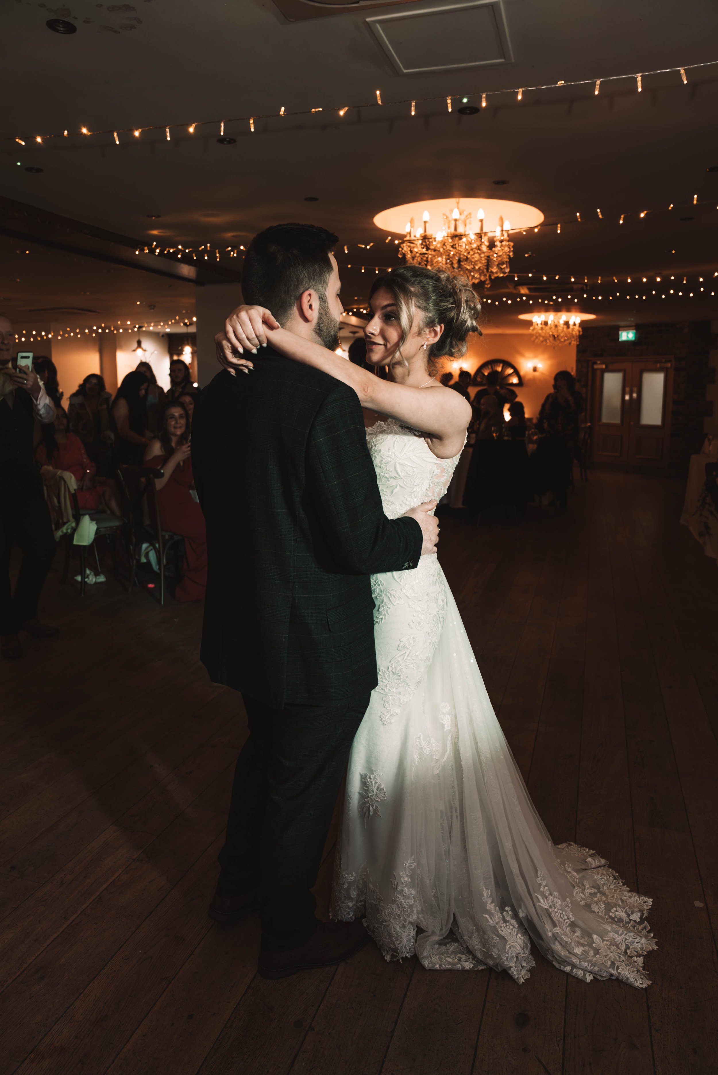 A bride and groom share their first dance at a wedding reception, surrounded by guests and warm lighting.
