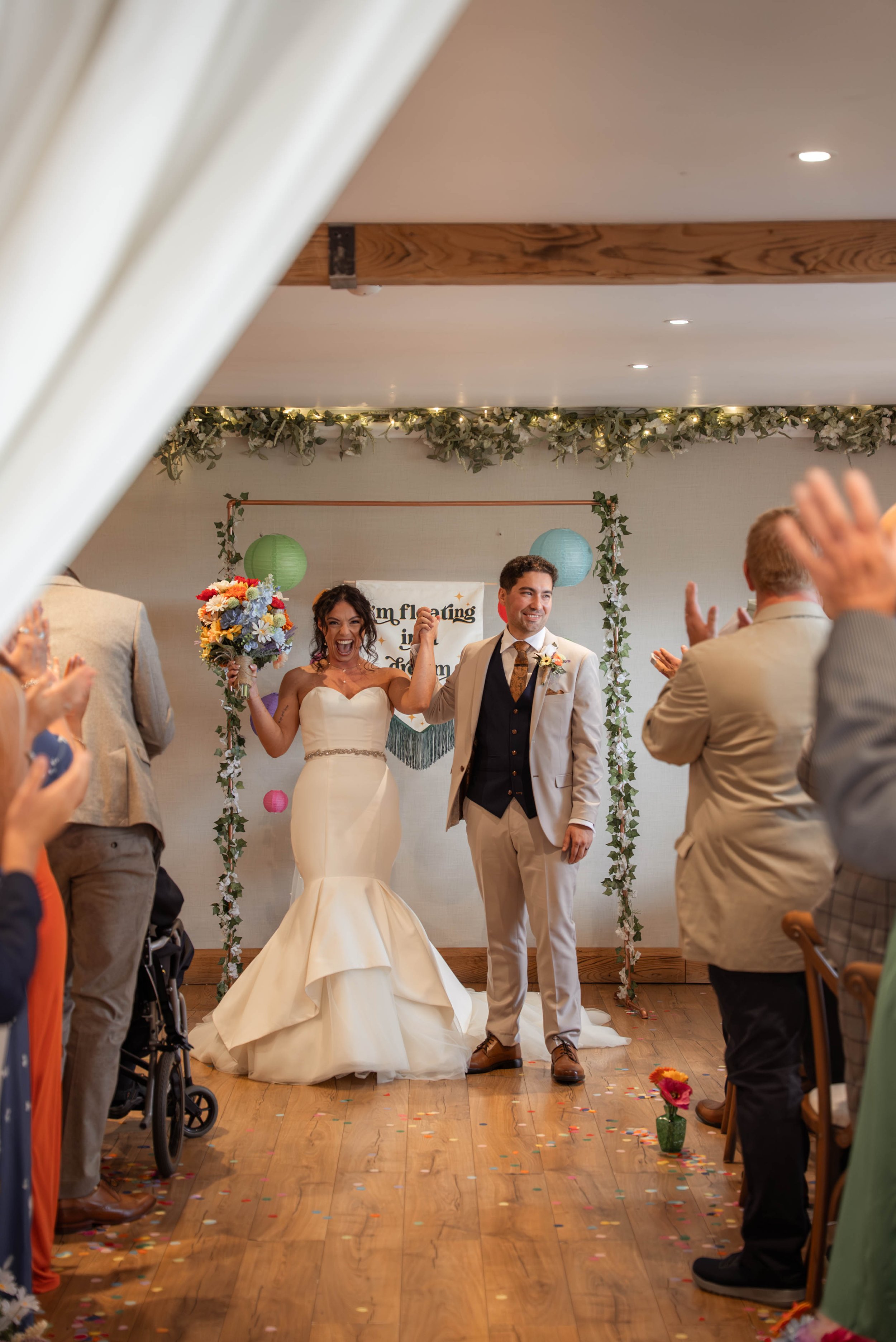 Newlywed couple celebrating at their wedding reception, standing in front of a decorated wall, with guests applauding around them. The bride is holding a colorful bouquet and appears joyful, while the groom is smiling, dressed in a light suit with a 