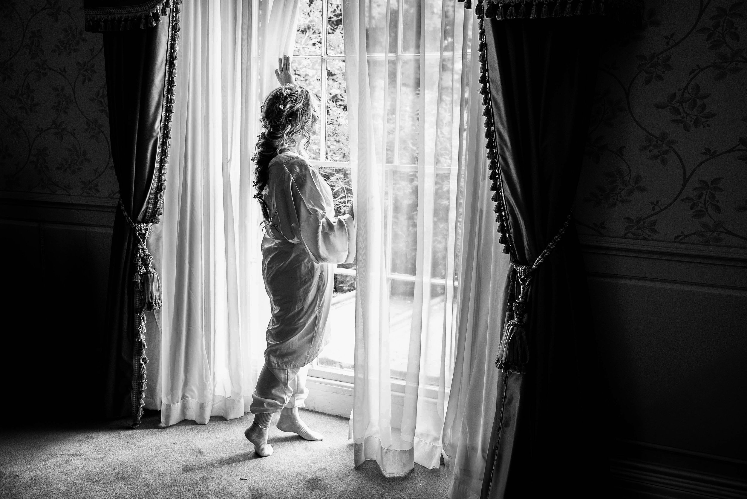 A woman with wavy hair wearing pajamas looking out a window with curtains, in a room with patterned wallpaper and carpet, black and white photograph.