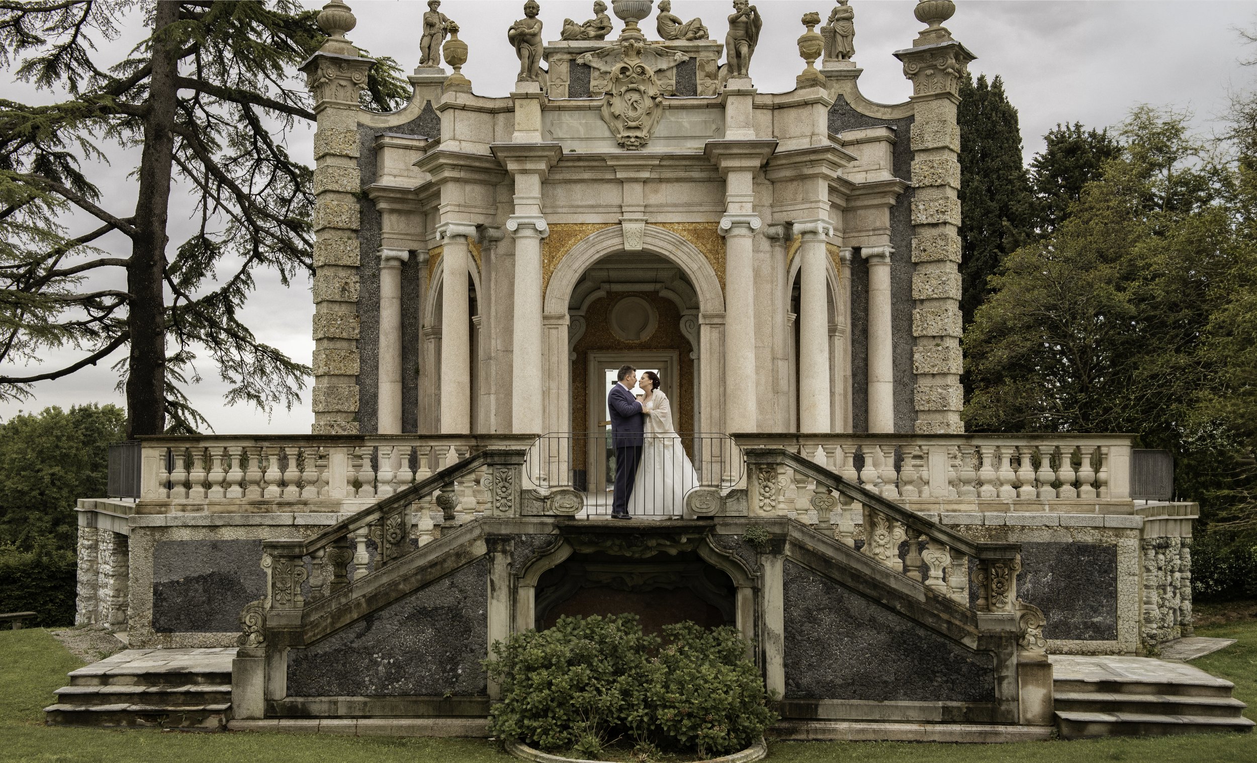 A couple in wedding attire standing on a balcony of an ornate, historic stone building, surrounded by trees with an overcast sky overhead.