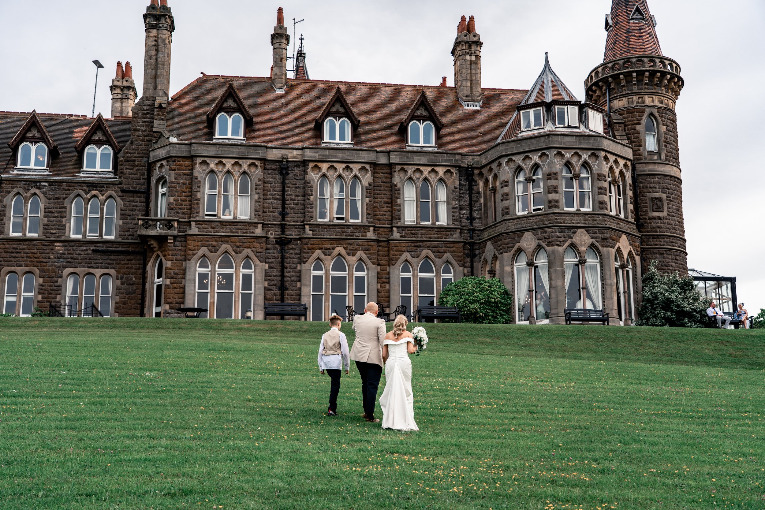 A bride and groom walking on a lawn towards a large historic stone building, with guests seated on benches and people standing nearby.