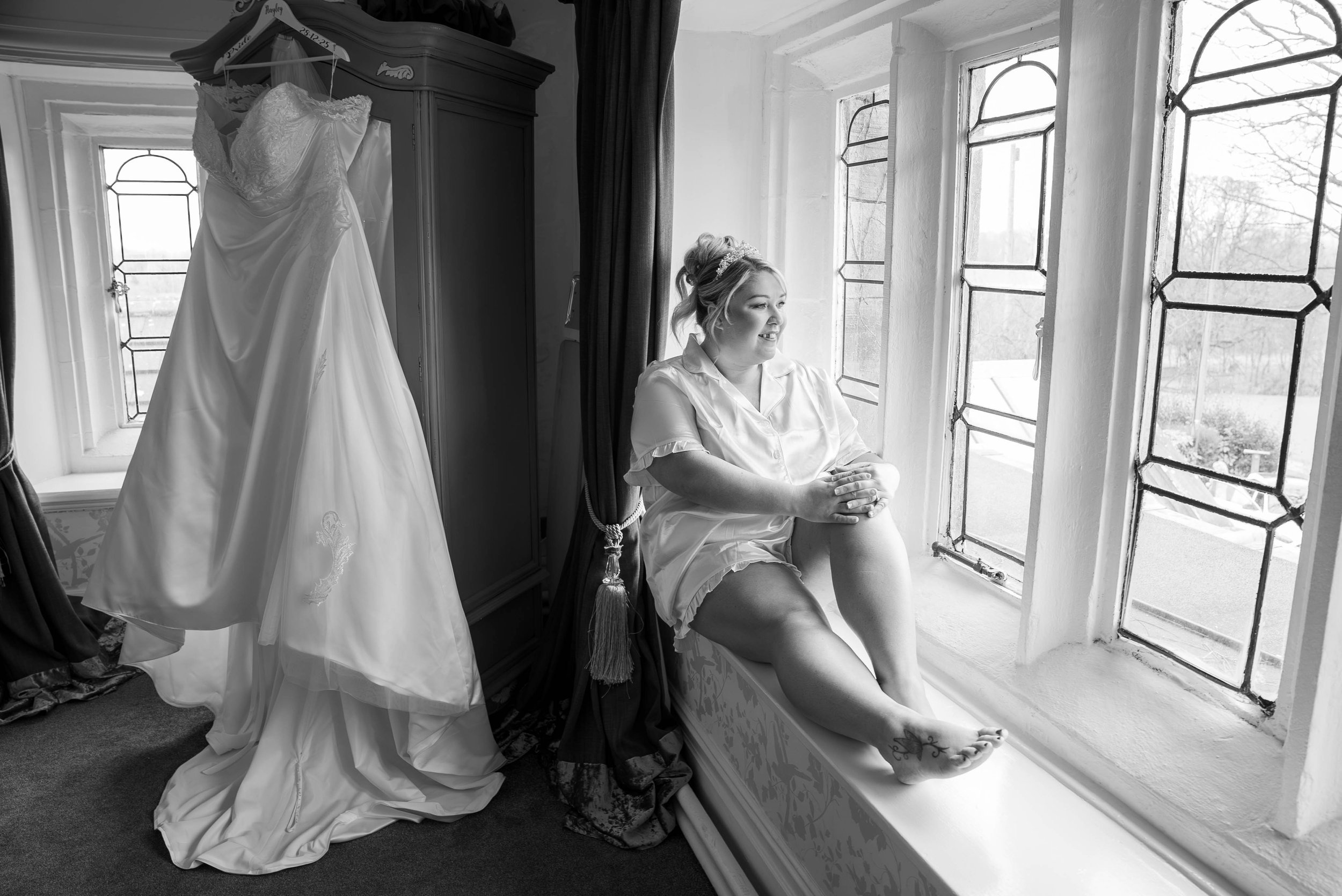 A woman sitting by a large window, smiling, dressed in light-colored clothes, with a wedding dress hanging nearby in a room with ornate furnishings.