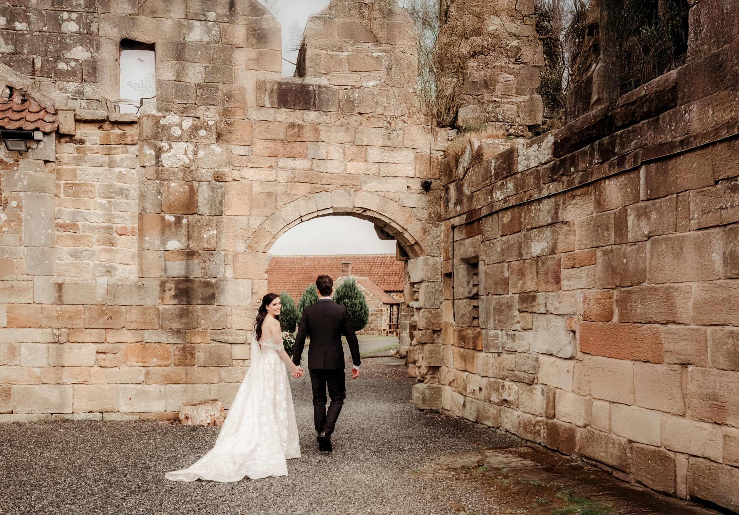 A bride and groom holding hands and walking through an old stone archway outdoors in a historical setting, with the bride in a white wedding gown and the groom in a black suit.