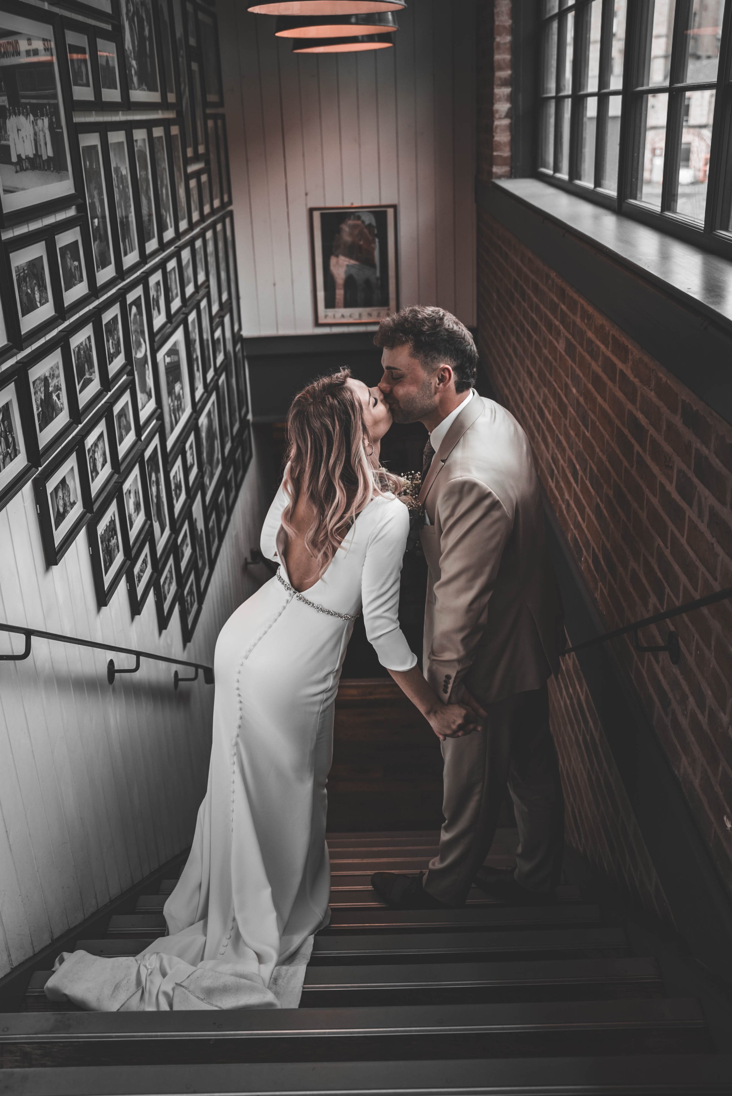A couple in wedding attire sharing a kiss on a staircase inside a building with framed photos on the wall.