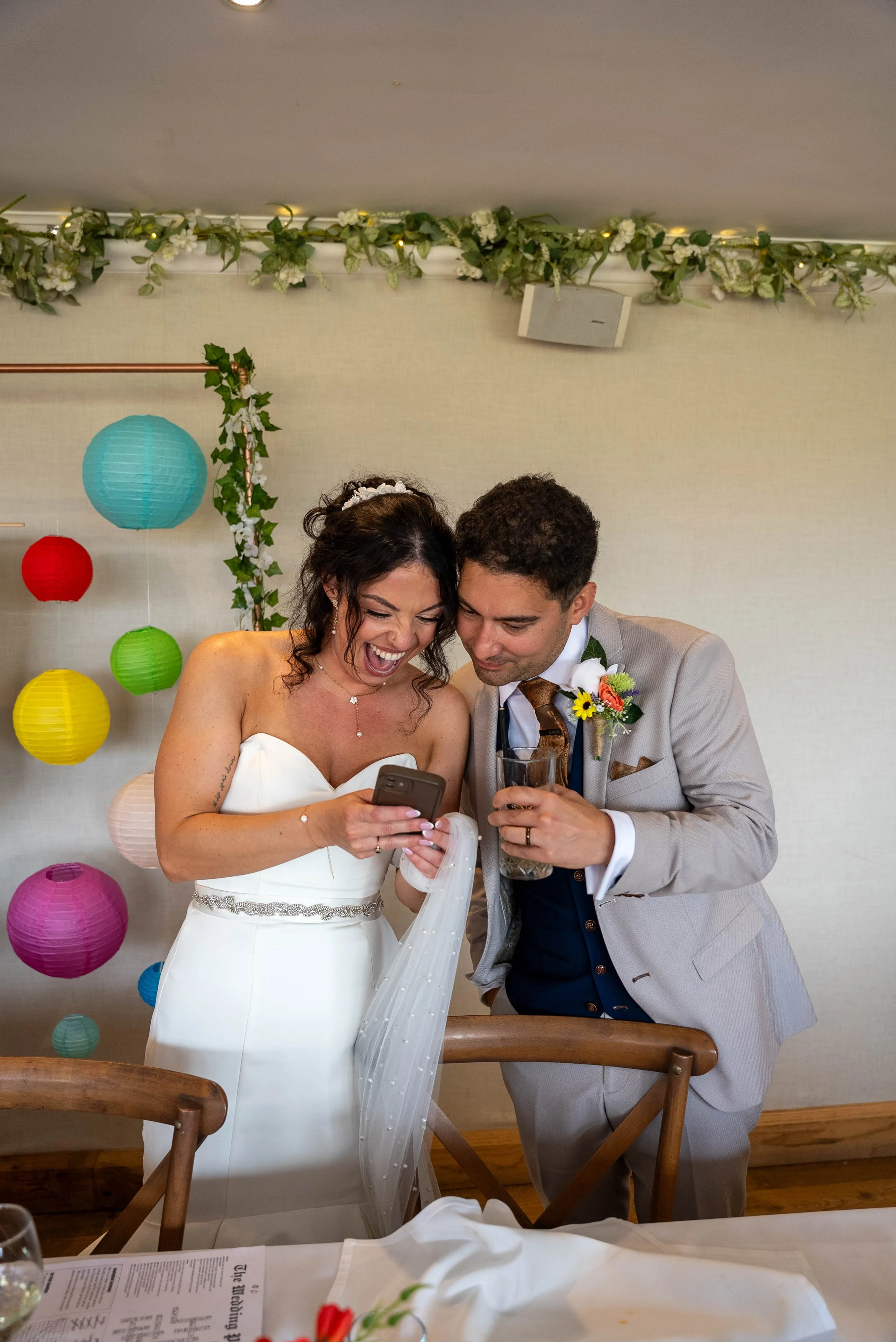 Bride and groom watching something on a phone at wedding reception, with colorful paper lanterns and floral decorations in the background.