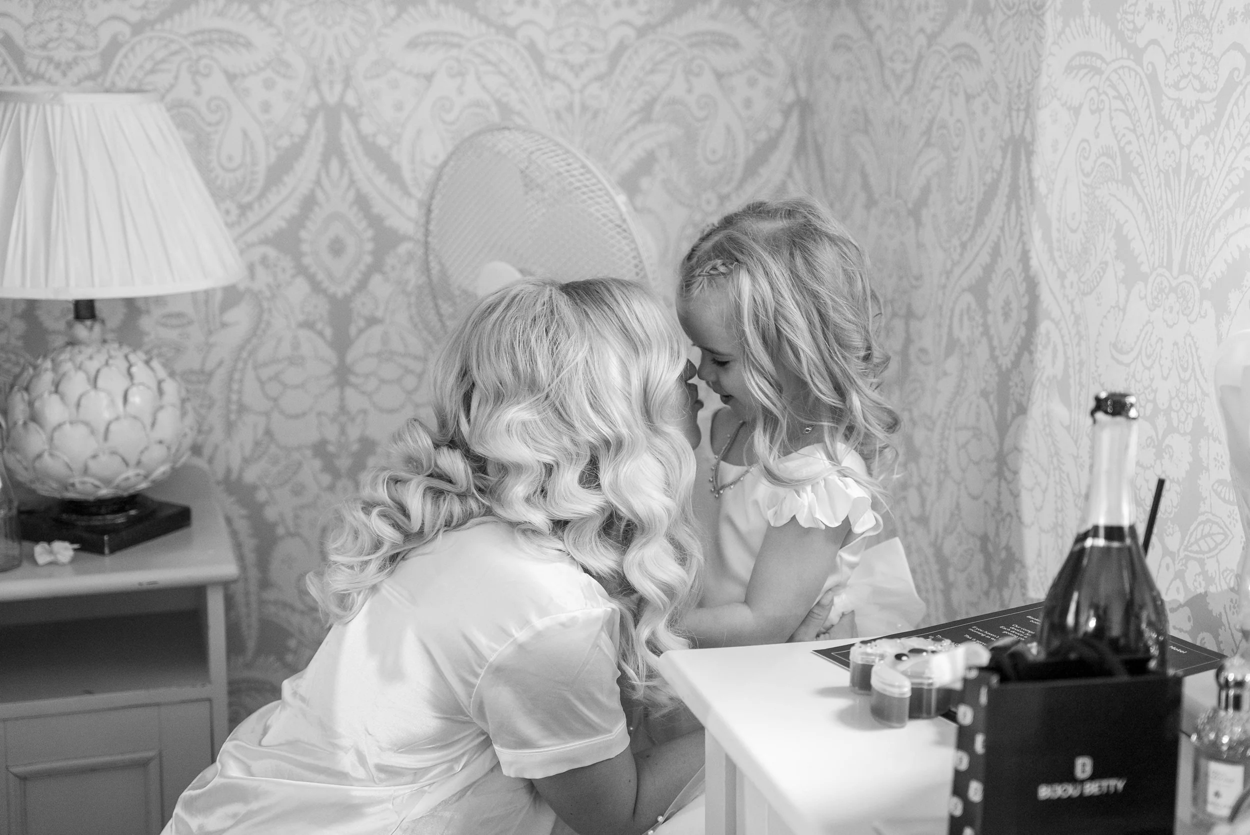 A woman and a young girl with curly hair touching foreheads in a room with vintage wallpaper, a table with makeup items, a lamp, and a bun hairstyle mirror.