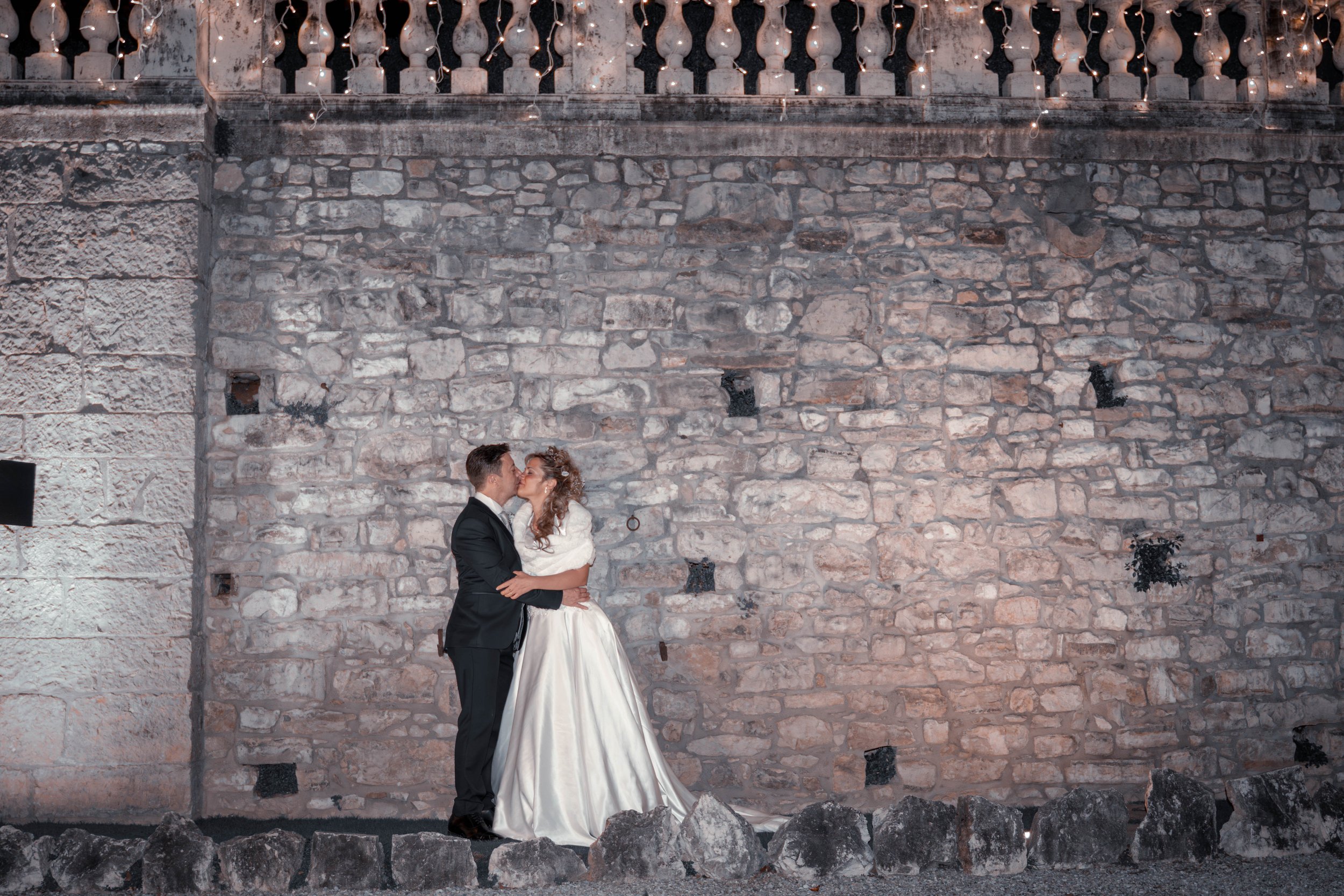 A bride and groom sharing a kiss in front of a stone wall with string lights hanging above.