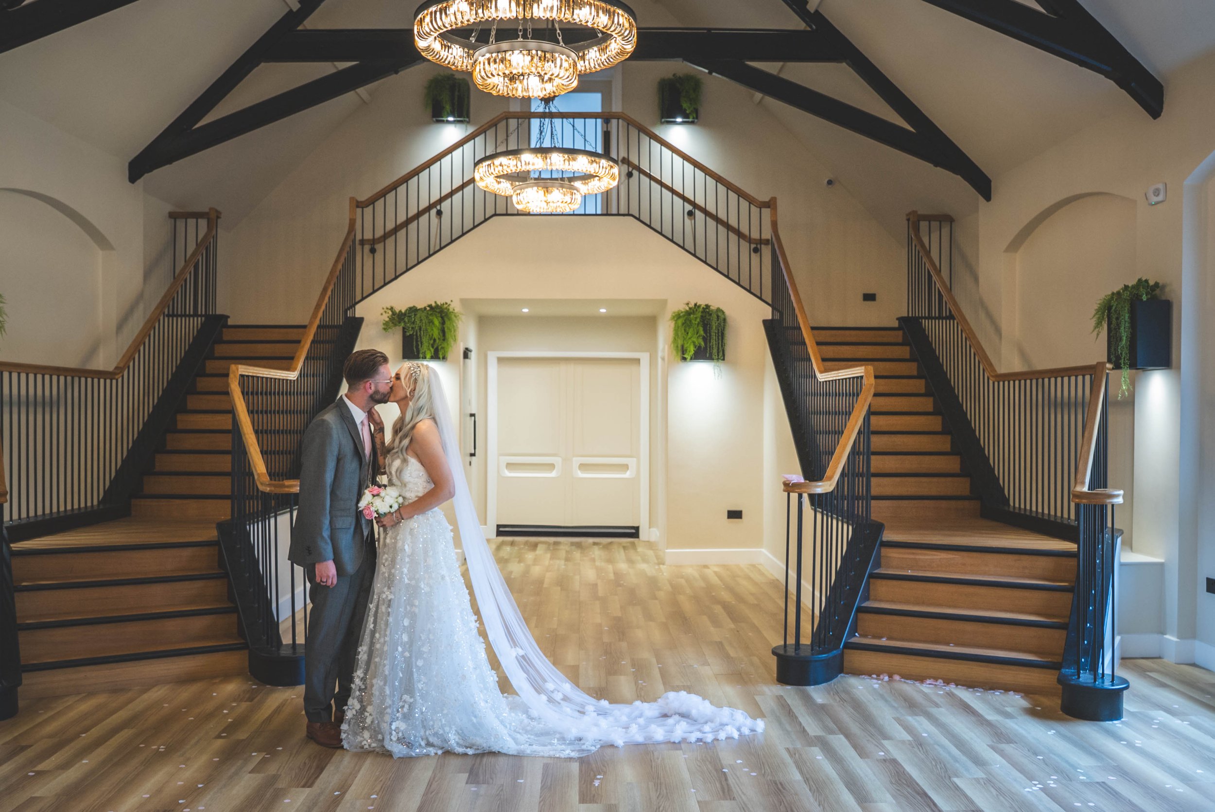 A bride and groom kissing in a decorated indoor venue with a staircase, chandeliers, and plant decorations.