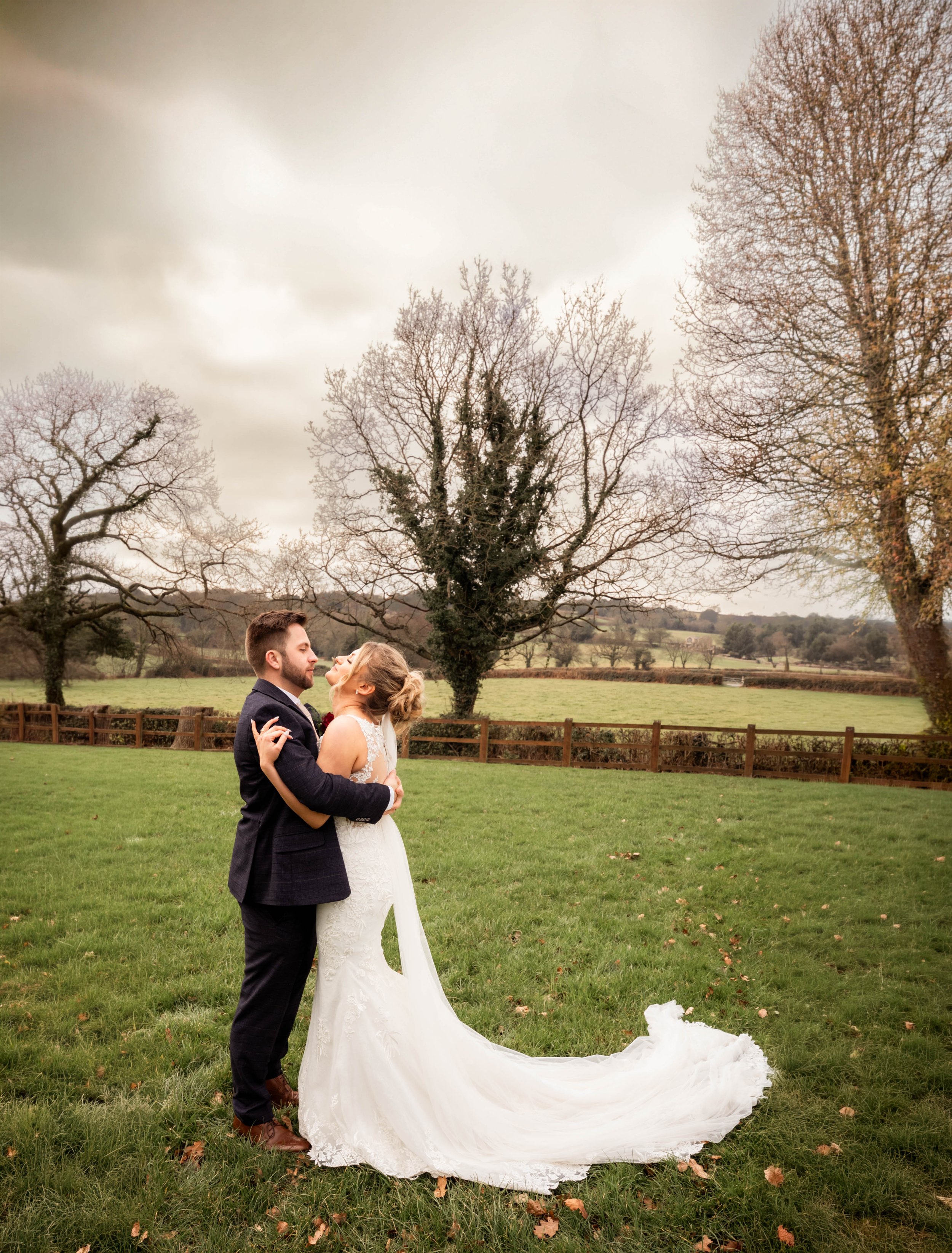 A bride and groom embracing outdoors on a cloudy day, with trees and fields in the background.
